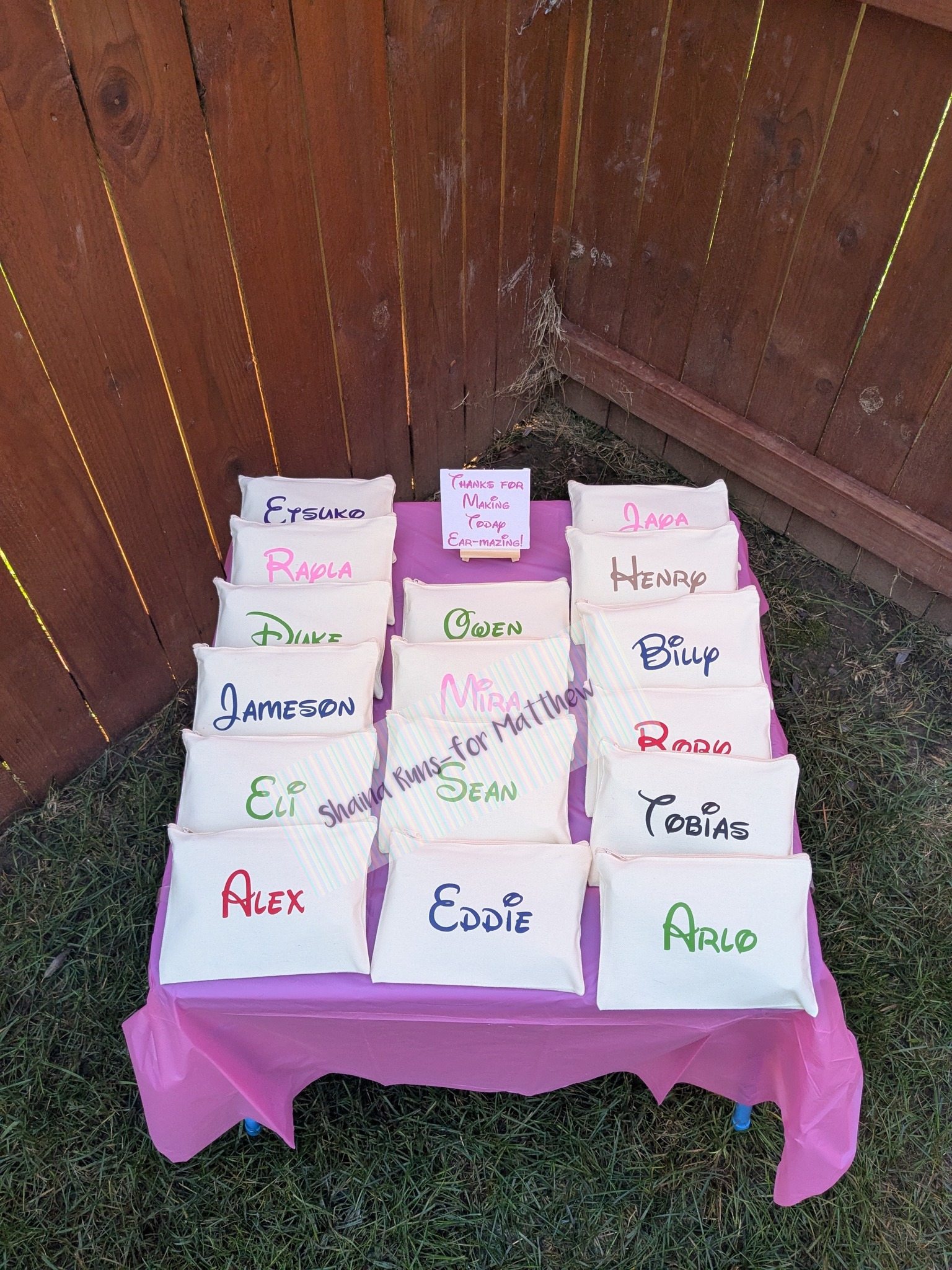 Display of personalized canvas favor bags, each featuring a guest's name, arranged on a pink table.