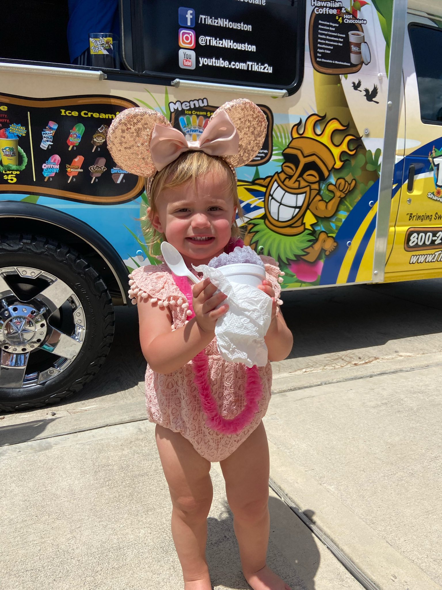 Toddler wearing mouse ears and a pink outfit holding a shave ice treat in front of a Tikiz ice cream truck.