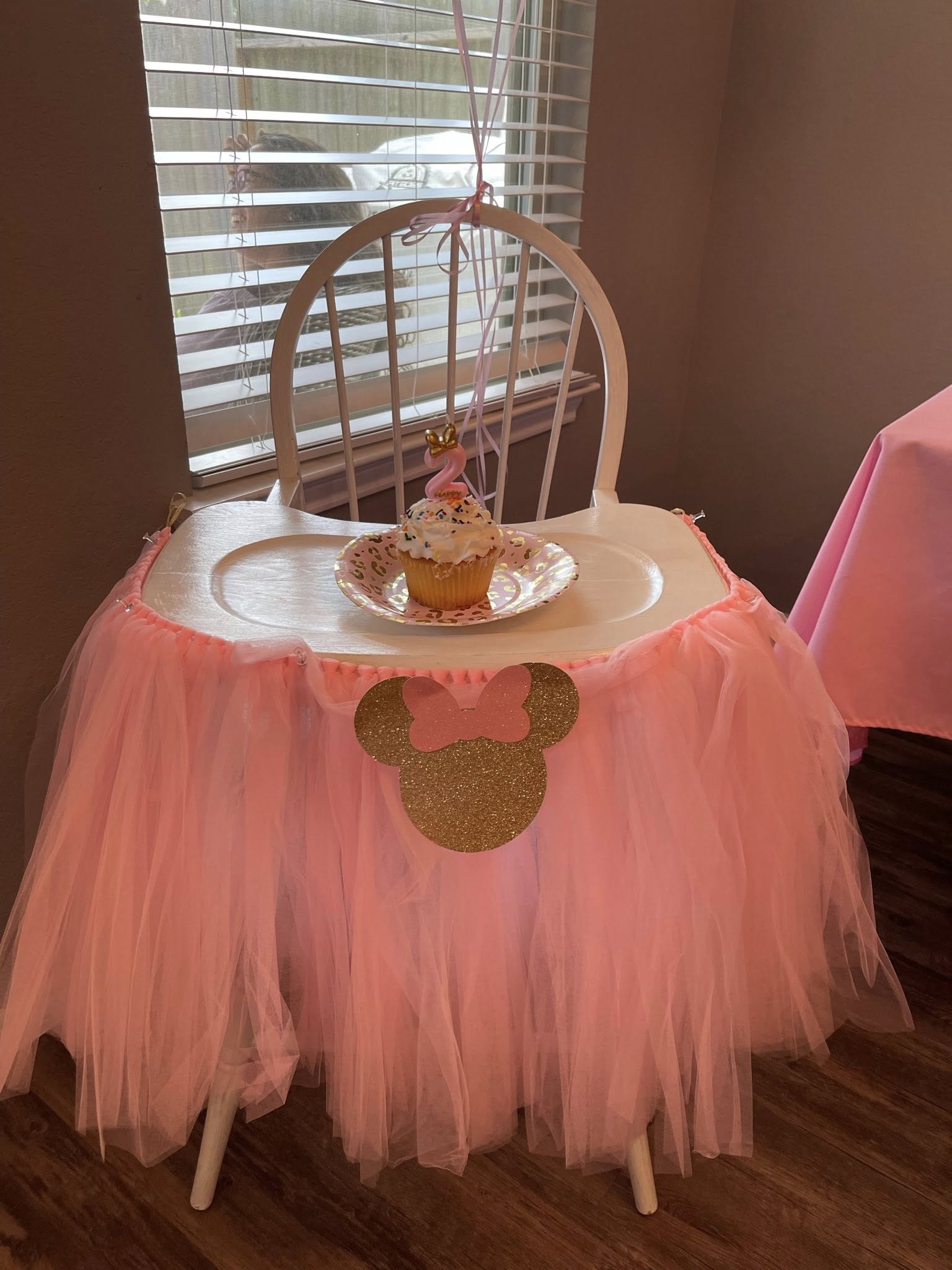 White high chair decorated with a pink tulle skirt and a gold Minnie Mouse ear cutout holding a cupcake with a number 2 candle.