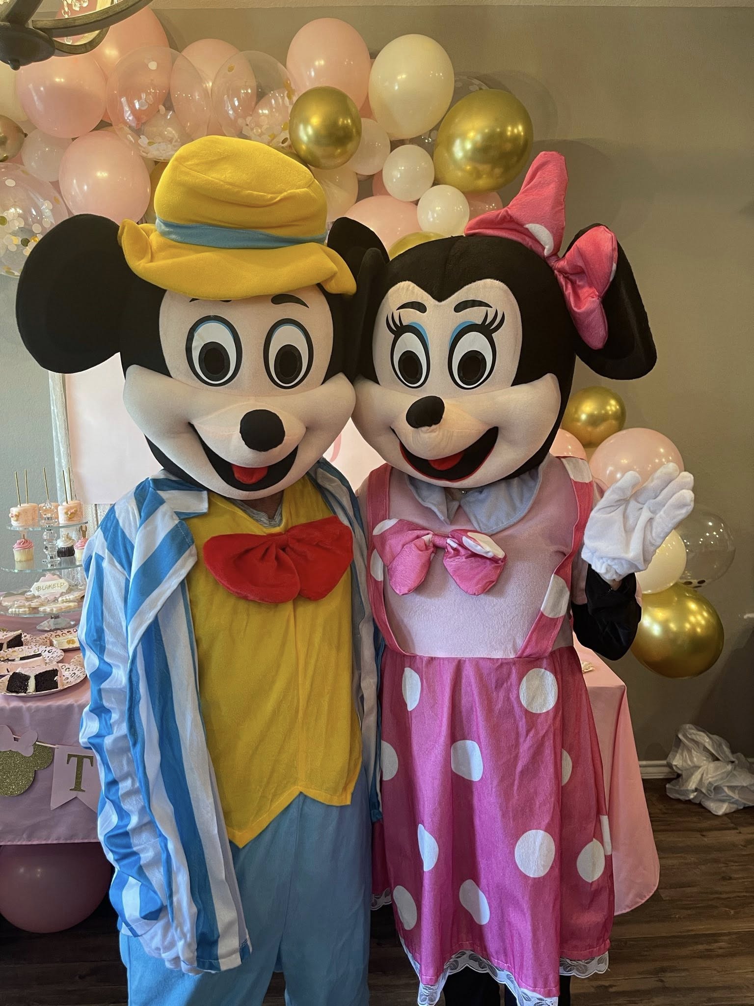 Mickey and Minnie Mouse mascot characters posing at a party with pink and gold balloon decorations.