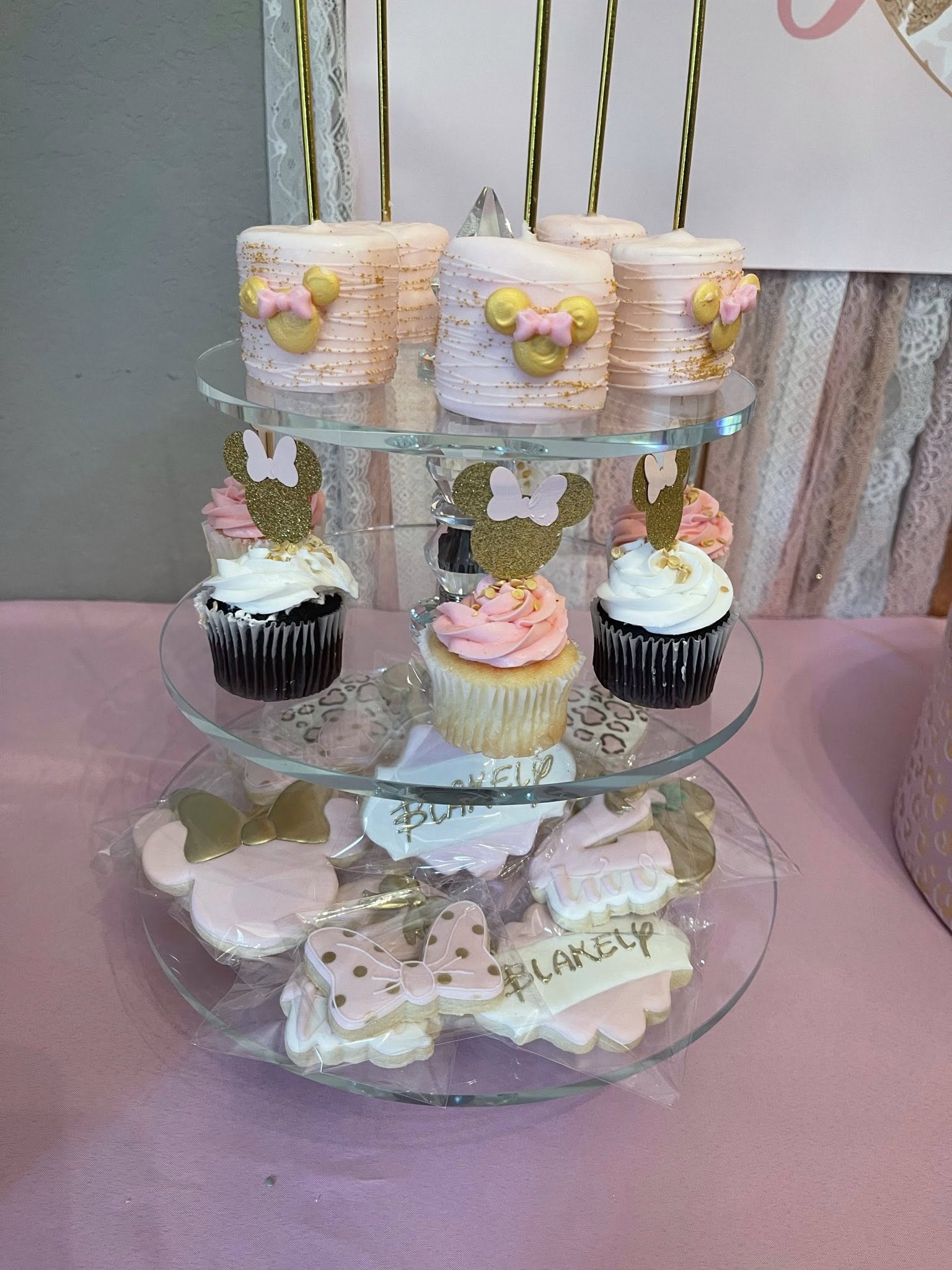Three-tiered display stand featuring Minnie Mouse themed cupcakes, cake pops, and custom sugar cookies for a party.