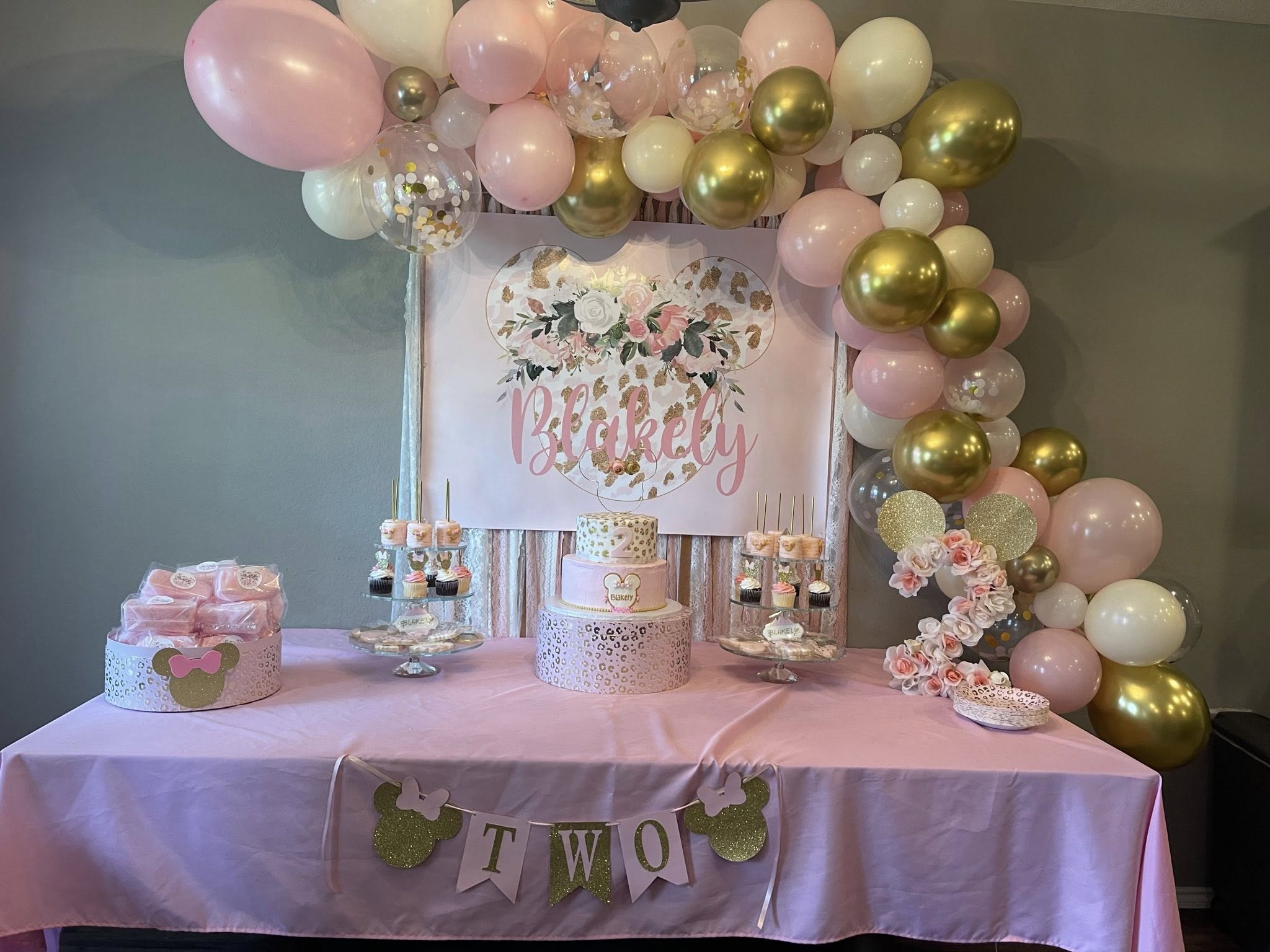 Table set up for a second birthday party with a pink and gold color theme, including a tiered cake and balloons.