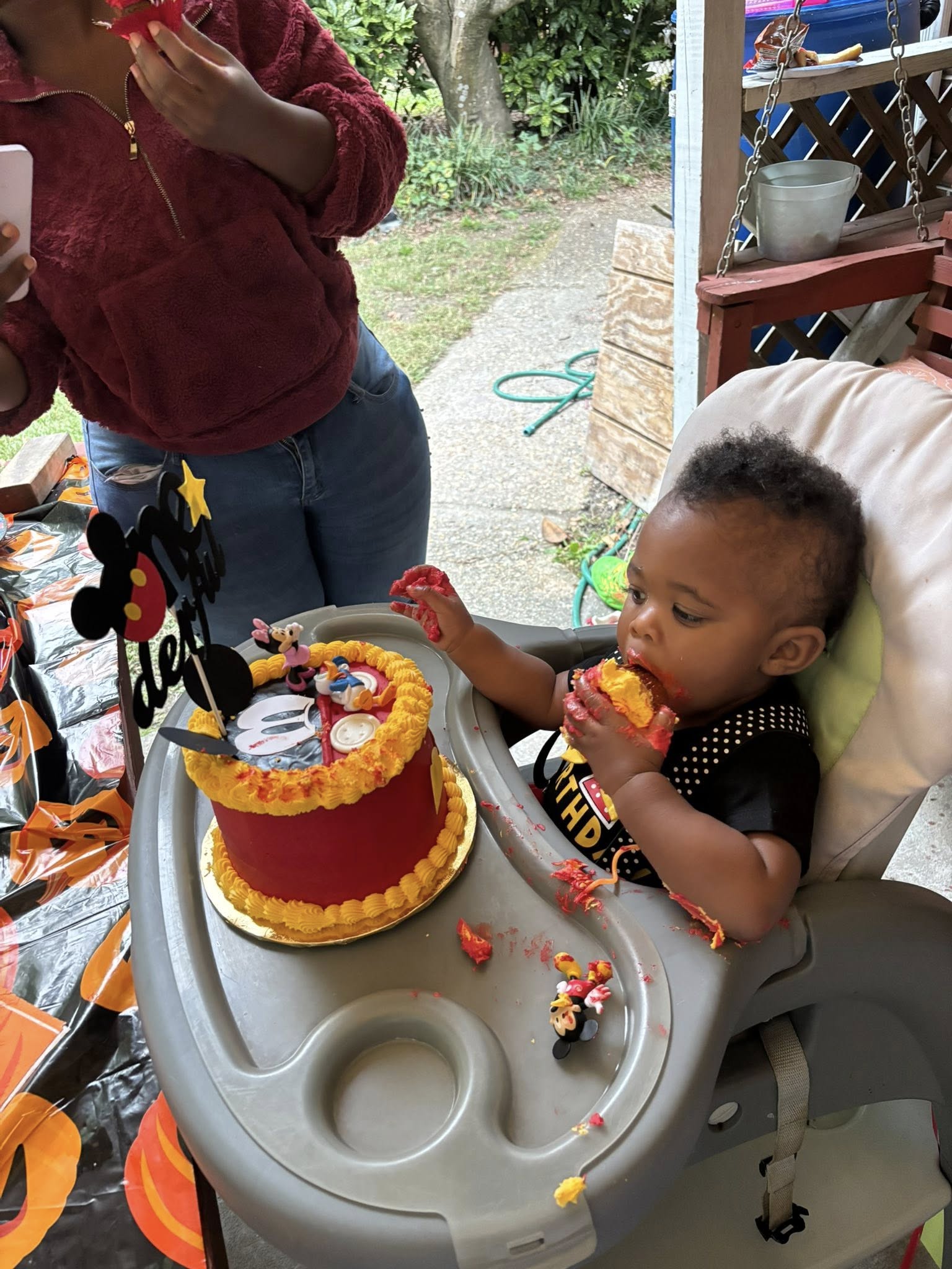 A baby eating a piece of red and yellow Mickey Mouse themed birthday cake in a high chair.