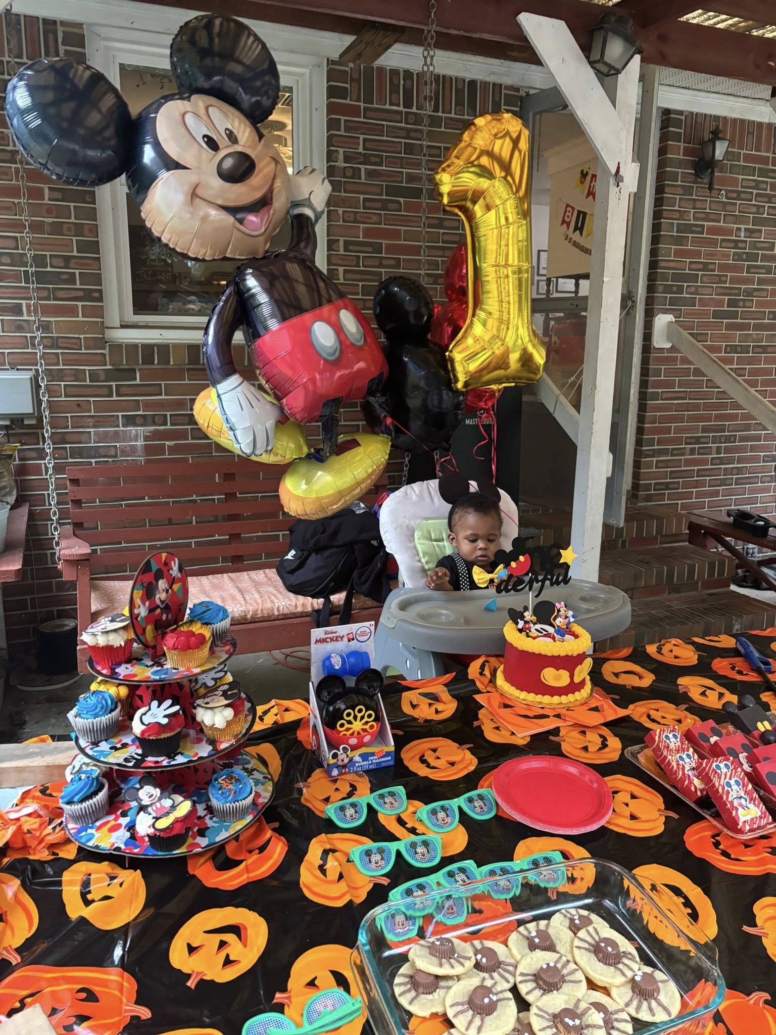 A one-year-old in a high chair at a mickey mouse themed first birthday party with balloons, cupcakes, and snacks.