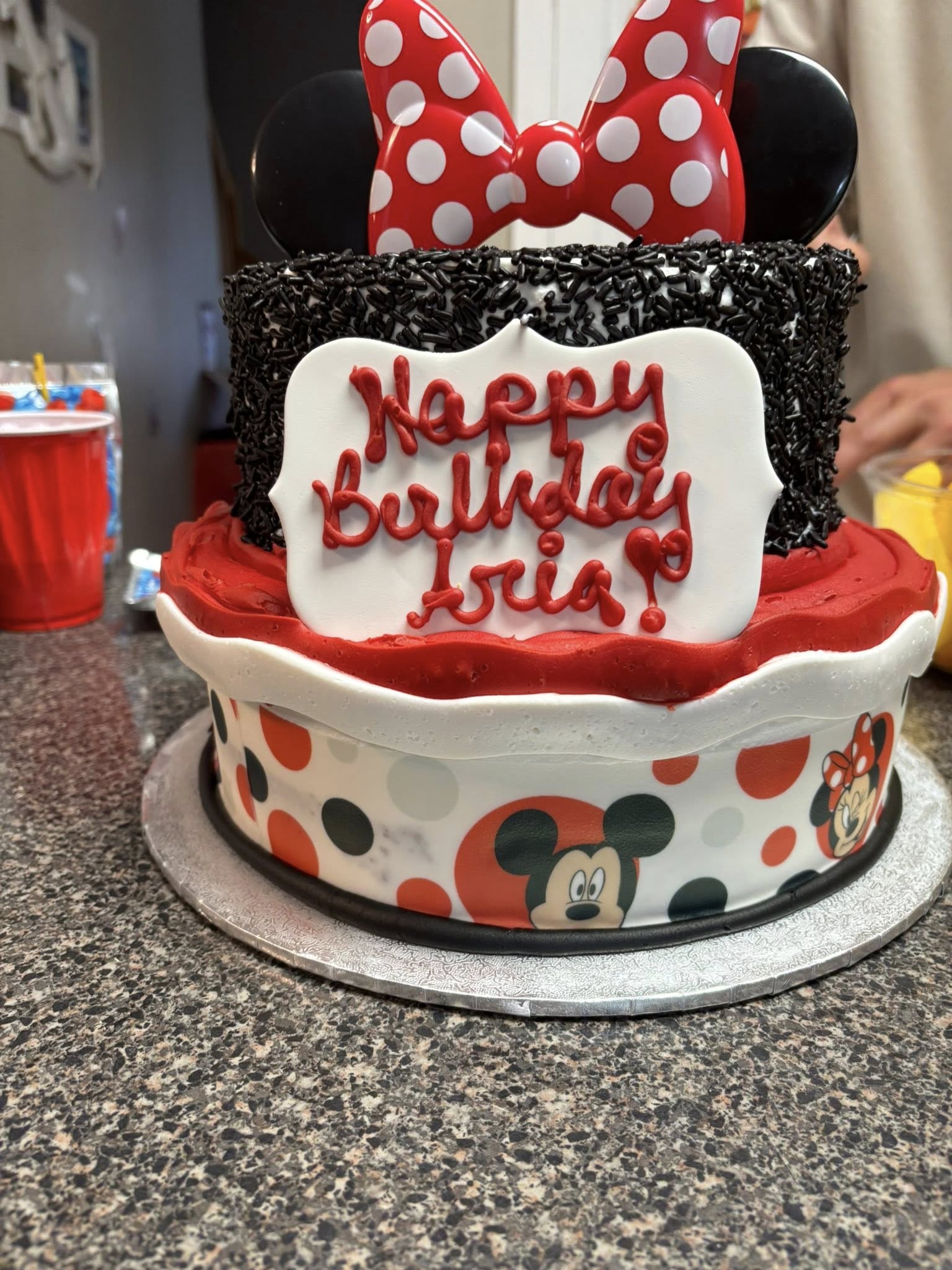 Two-tier Minnie Mouse themed birthday cake with red, white, and black decorations and Happy Birthday Aria inscription.