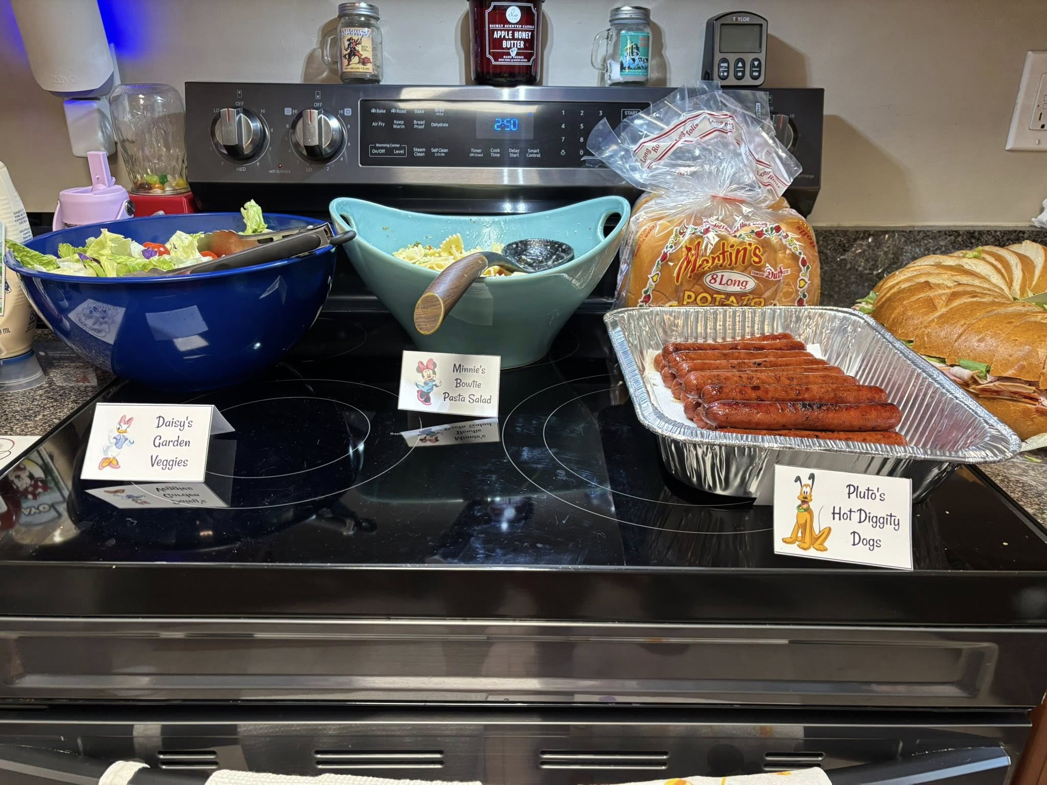 A Disney-themed food spread on a stove featuring bowls of garden salad, pasta salad, hot dogs, and sandwich rolls.