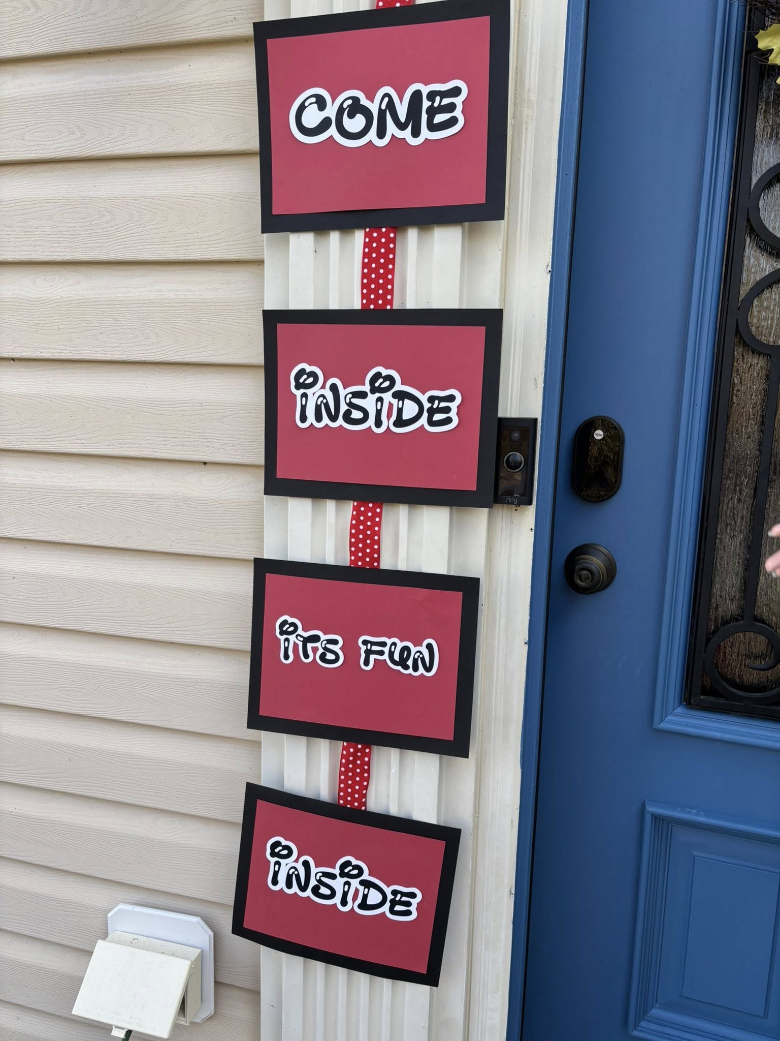 Set of four hanging signs on a red ribbon spelling out Come Inside It's Fun hanging next to a blue front door.