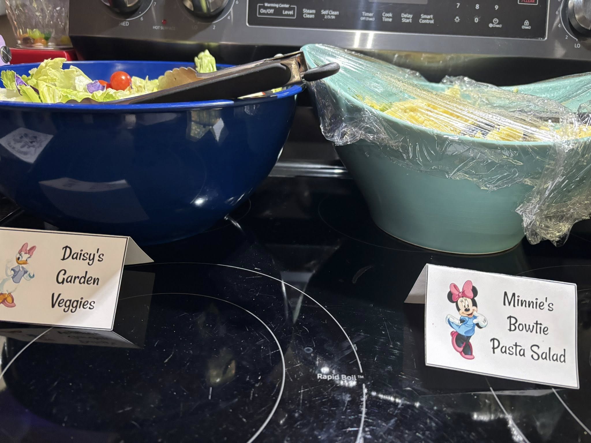 Daisy's Garden Veggies salad and Minnie's Bowtie Pasta Salad with character themed food labels on a stovetop.