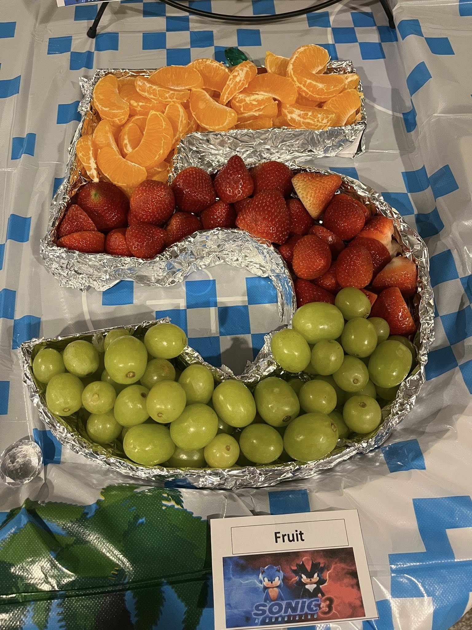Number 5 shaped fruit platter with orange slices, strawberries, and green grapes on a blue and white checkered cloth