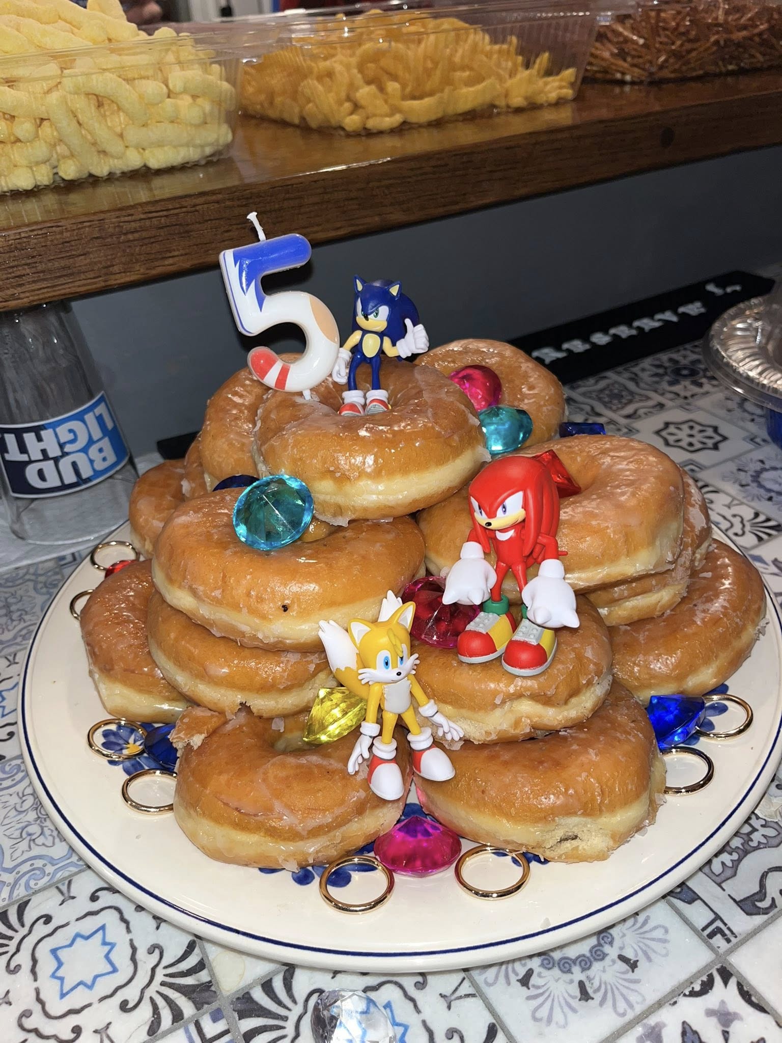 Stack of glazed donuts decorated with a number 5 candle and Sonic the Hedgehog character figurines and plastic gems.