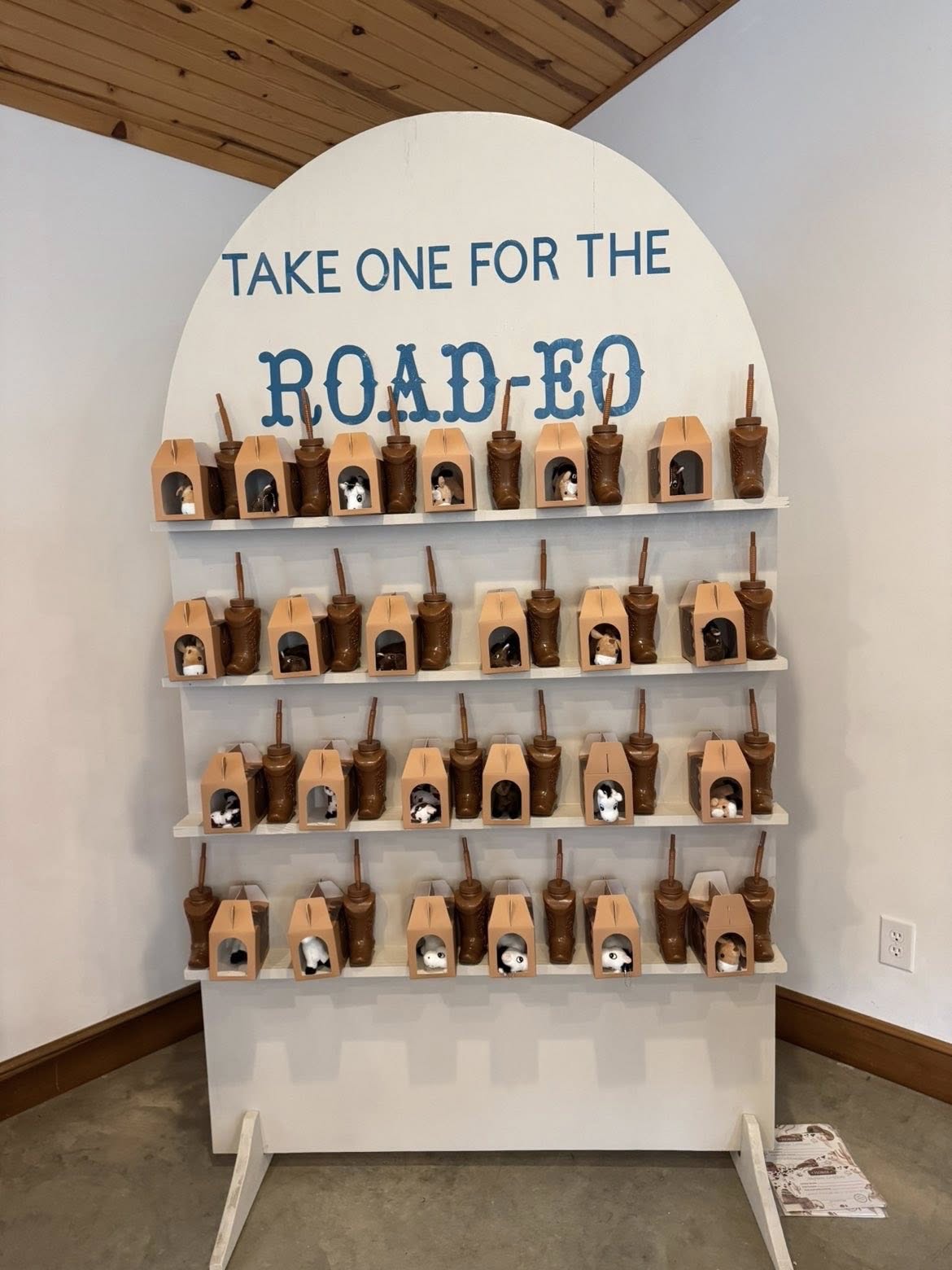 Display rack filled with souvenir boot-shaped cups featuring small mascot figures, with a sign reading Take One for the Road-eo.