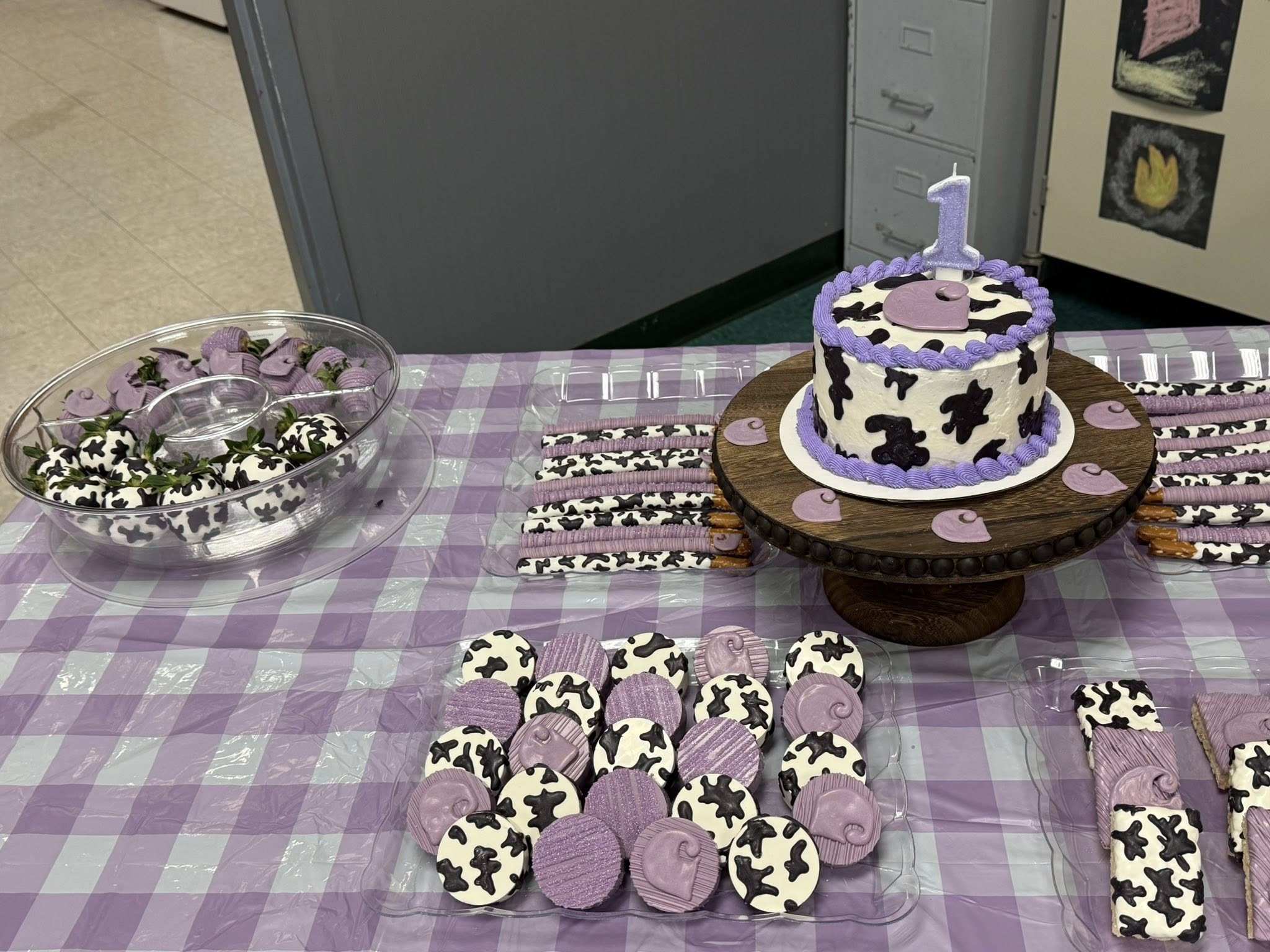 Table set with purple and black cow-print themed first birthday cake, chocolate-covered strawberries, and cookies.