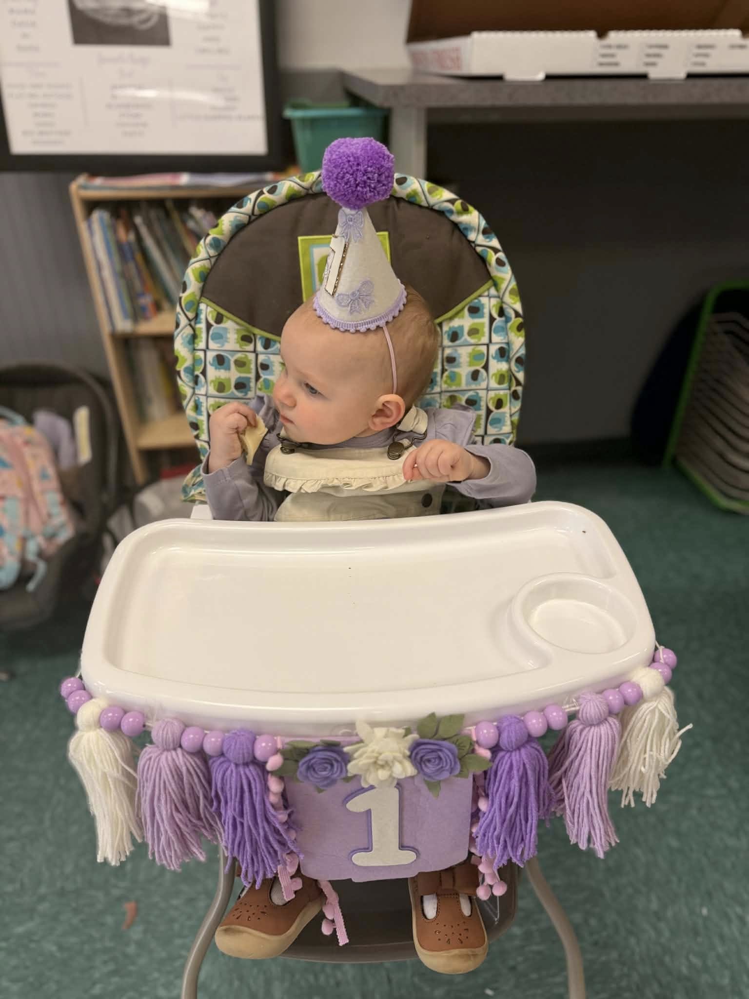 First birthday high chair banner in purple and white with yarn tassels and floral decorations