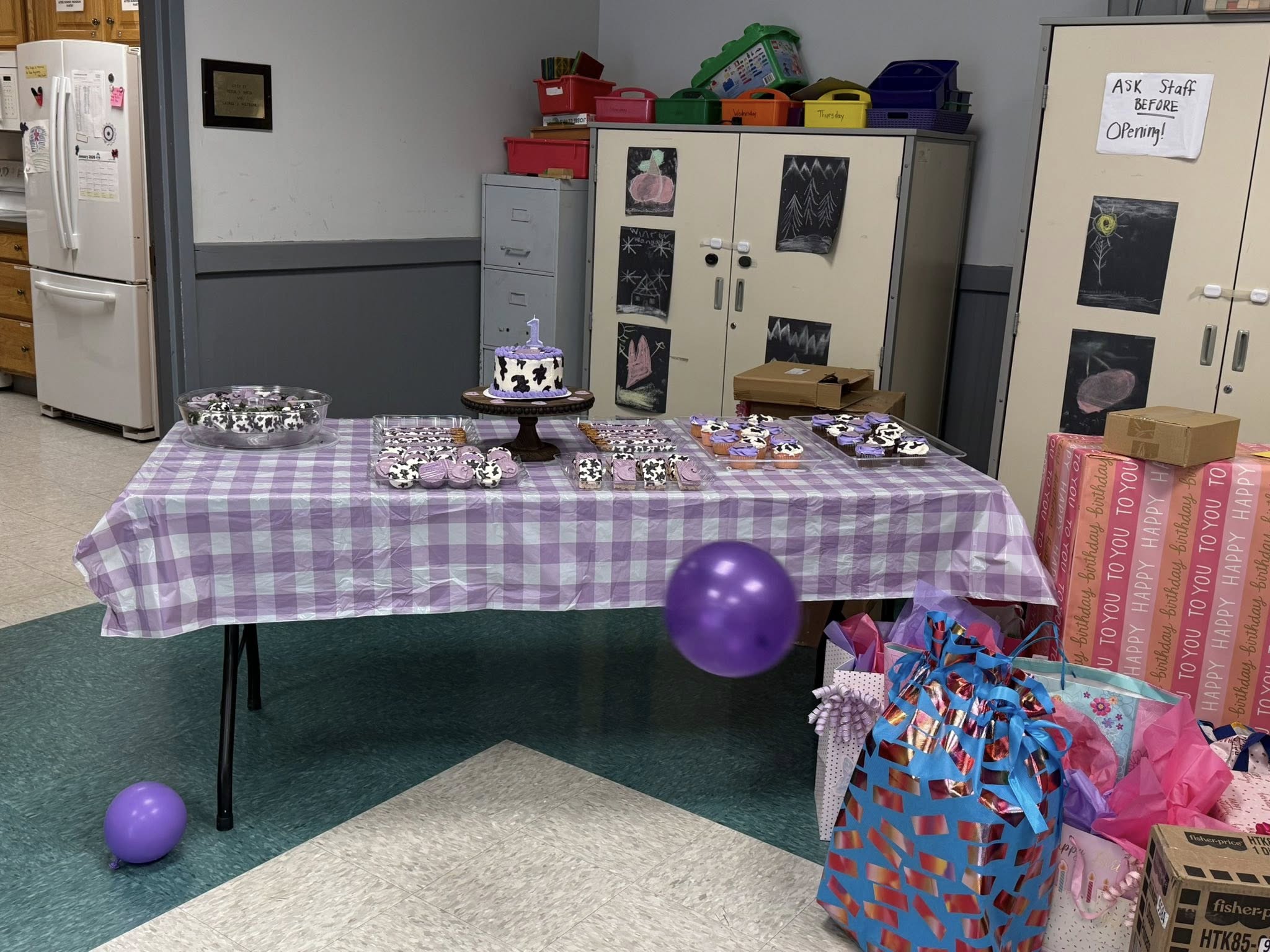 Birthday party table set with a cow-print cake and cupcakes on a purple checkered tablecloth.