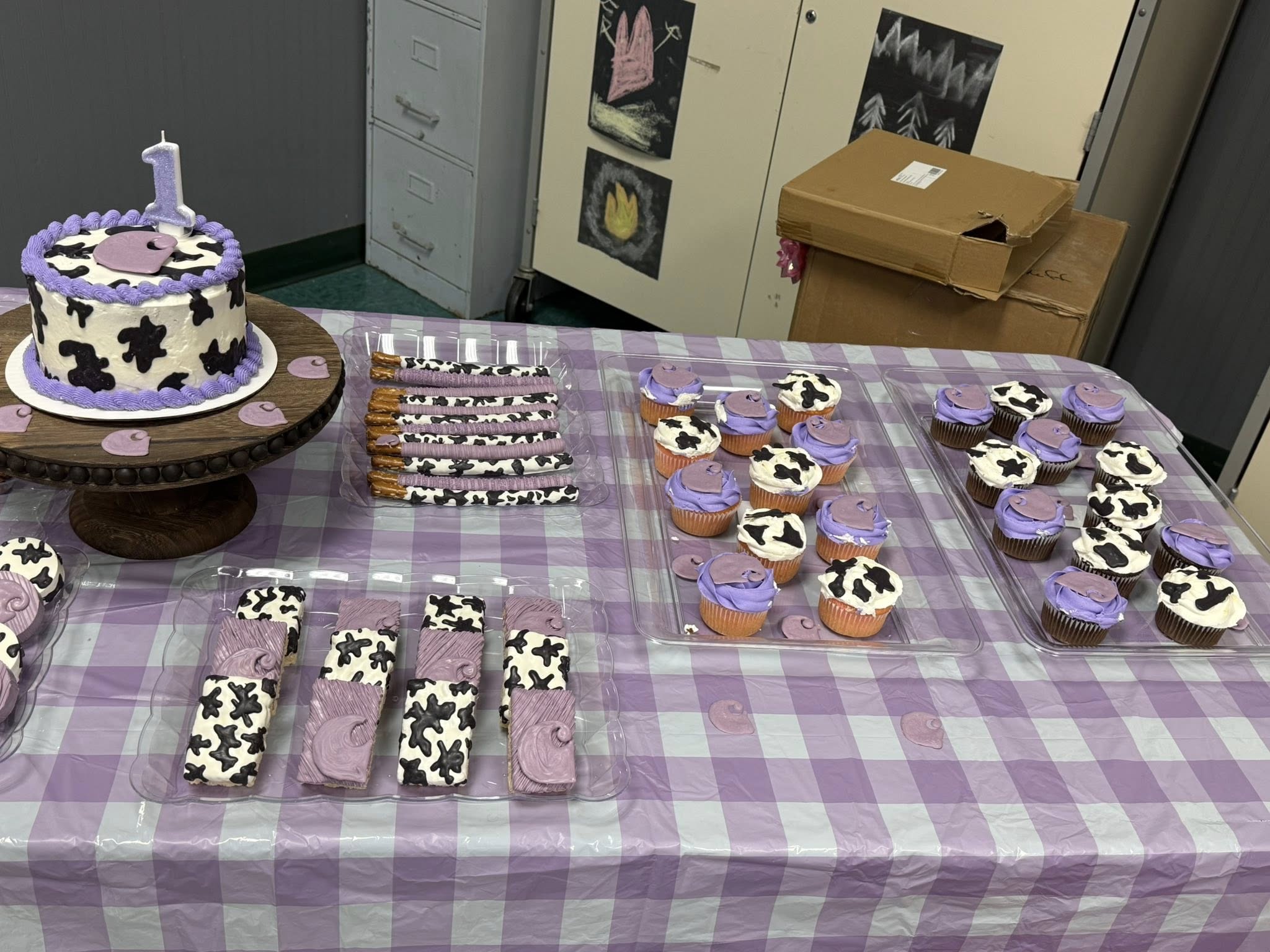 Cow print and purple 1st birthday party treats including a cake, cupcakes, and chocolate covered pretzel rods.