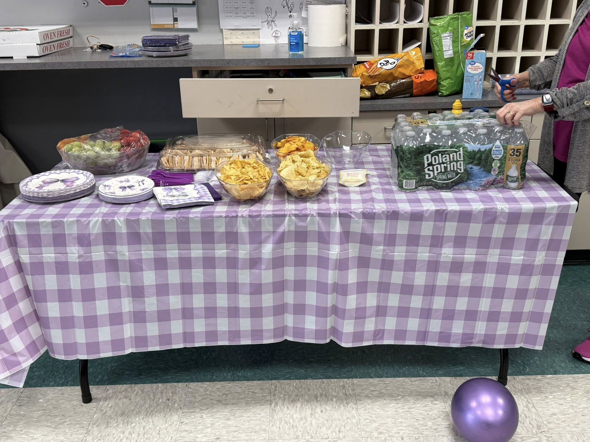 A table covered by a lavender checkered tablecloth, set for a party with snack bowls, finger sandwiches, fruit, and water bottles.