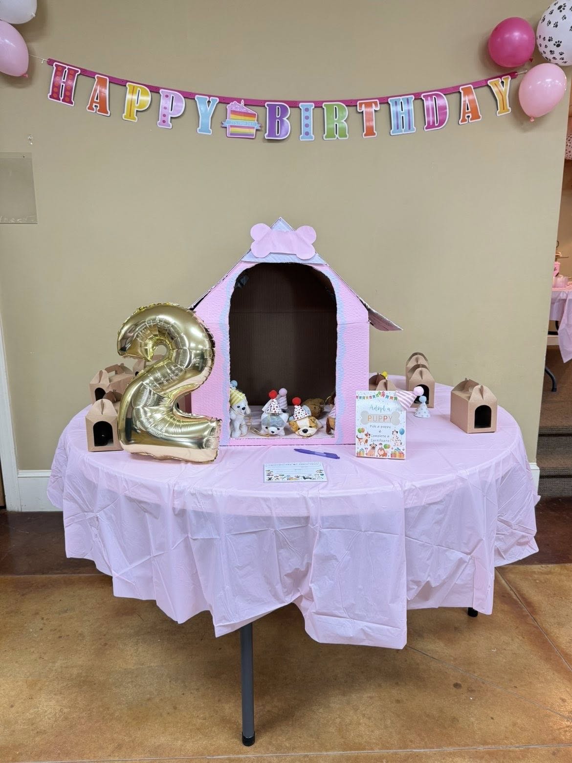Puppy adoption birthday party table with a pink DIY dog house, plush puppies, and a gold number two balloon.