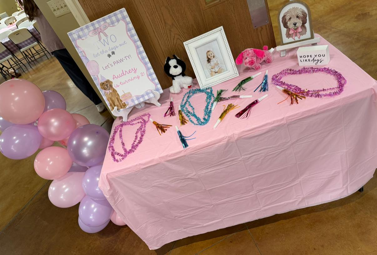 Table decorated for a girl's second birthday party with paw print sign, photos, and pink and purple party favors.