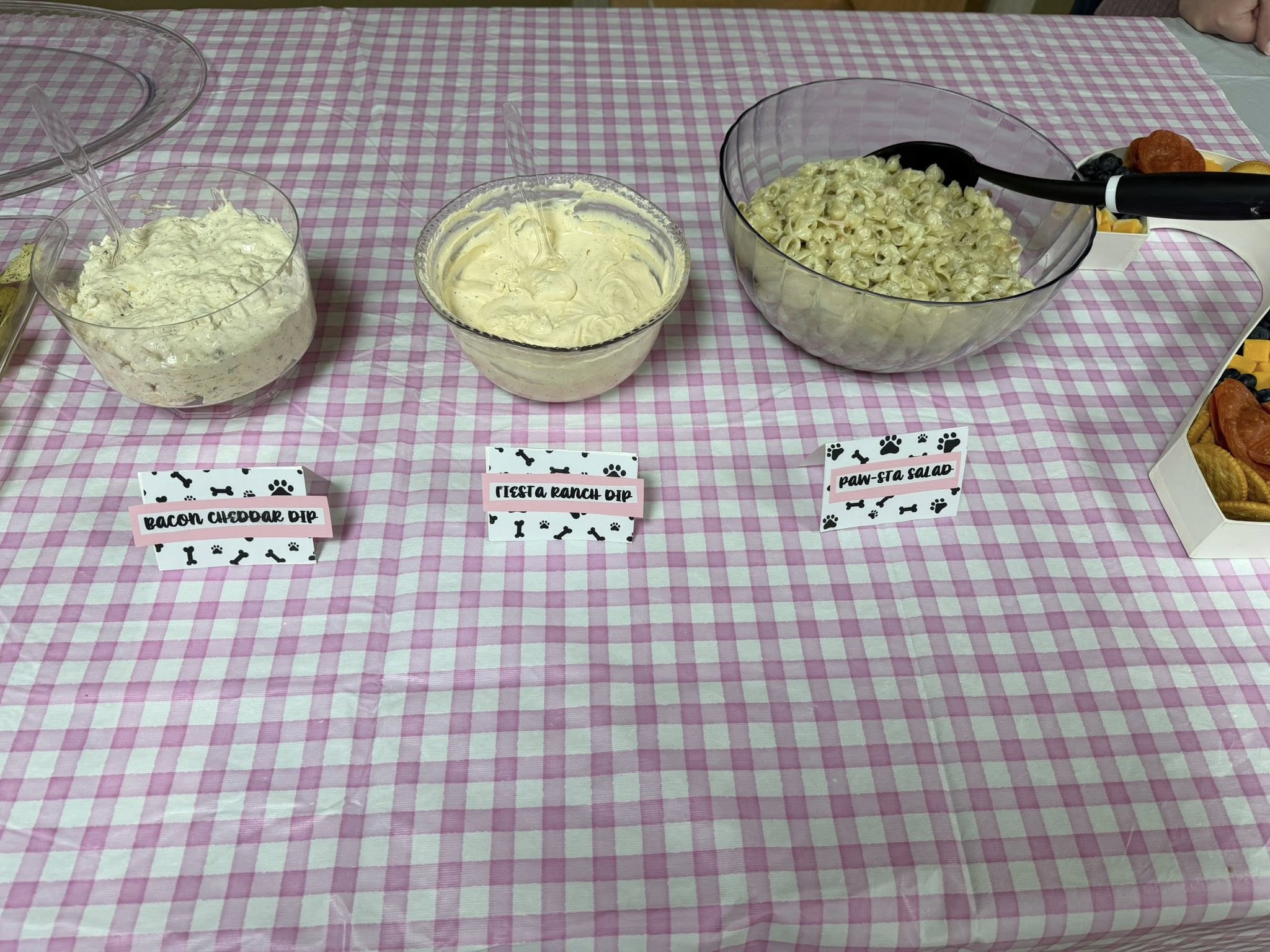 Three bowls of appetizers on a pink gingham tablecloth with labels reading Bacon Cheddar Dip, Fiesta Ranch Dip, and Paw-sta Salad.