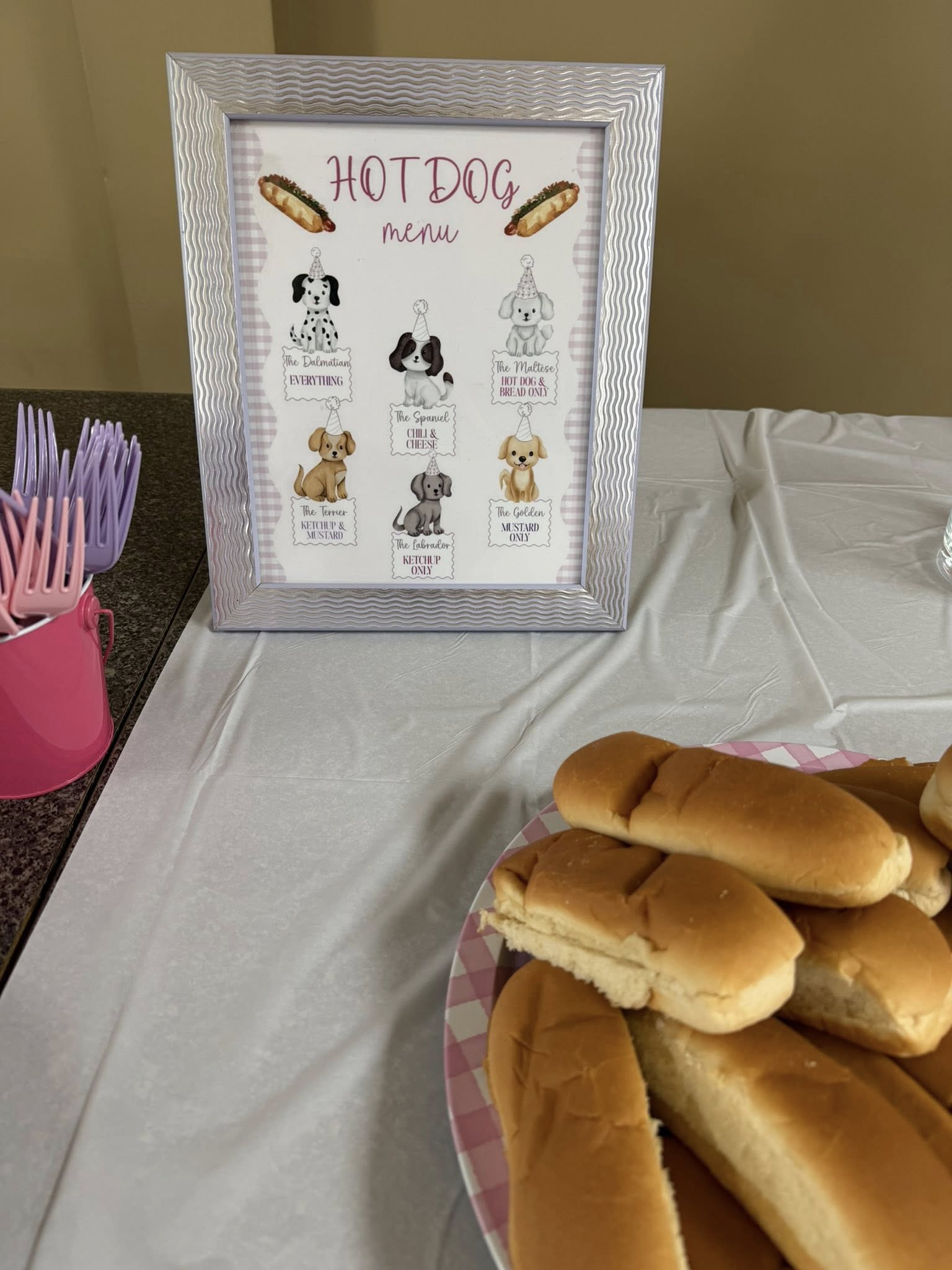 Table with a framed hot dog menu featuring dog illustrations and a plate of plain hot dog buns.