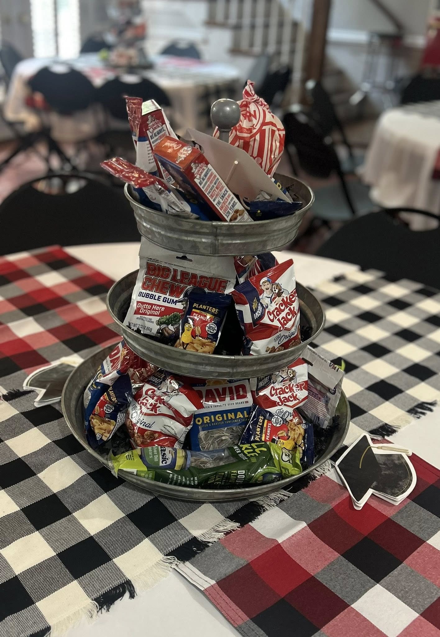 Three-tiered galvanized metal tray filled with assorted snack bags including Cracker Jack, Big League Chew, and peanuts.