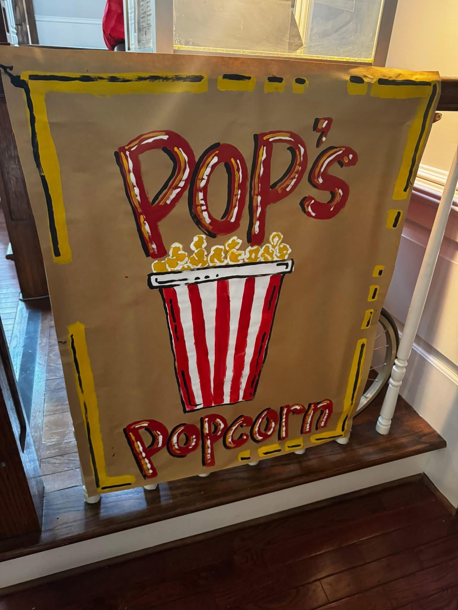 Hand-painted sign on brown paper reading Pop's Popcorn with a graphic of a red and white striped popcorn bucket.