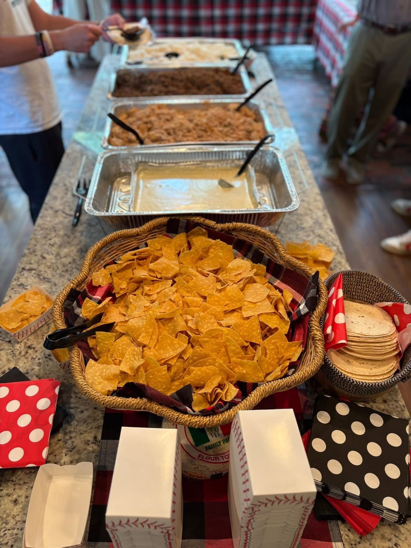 Taco bar setup featuring baskets of tortilla chips, flour tortillas, and several catering trays of meat and queso.