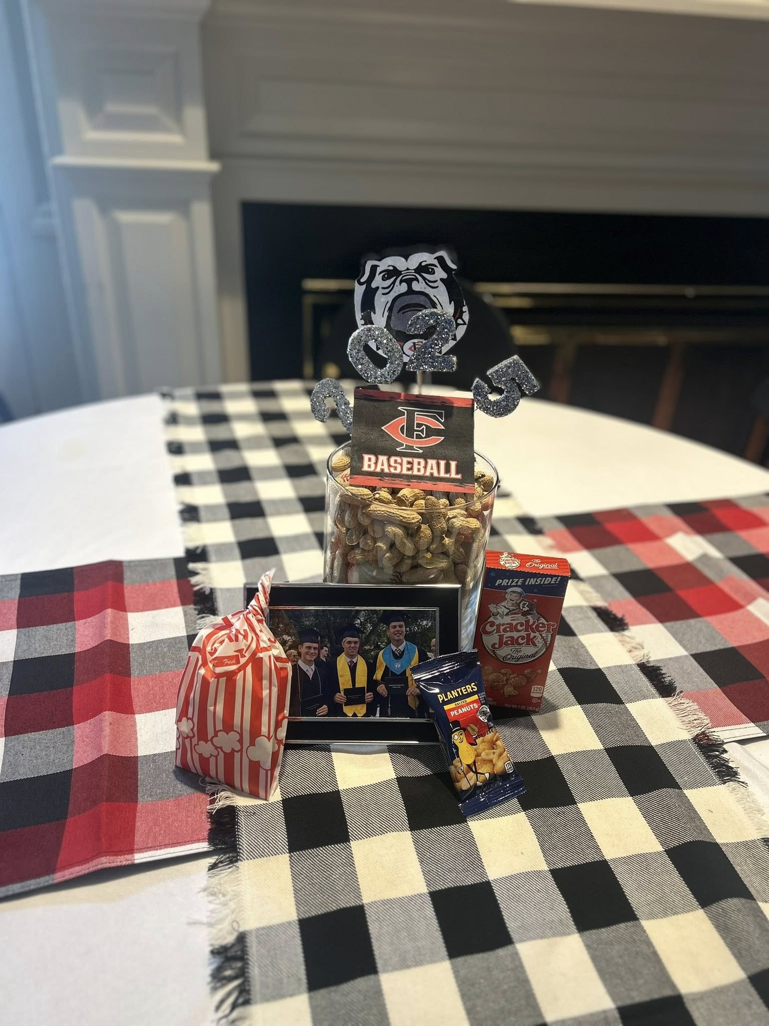 Table centerpiece featuring a graduation 2025 sign, a jar of peanuts, a photo frame, and classic ballpark snacks.