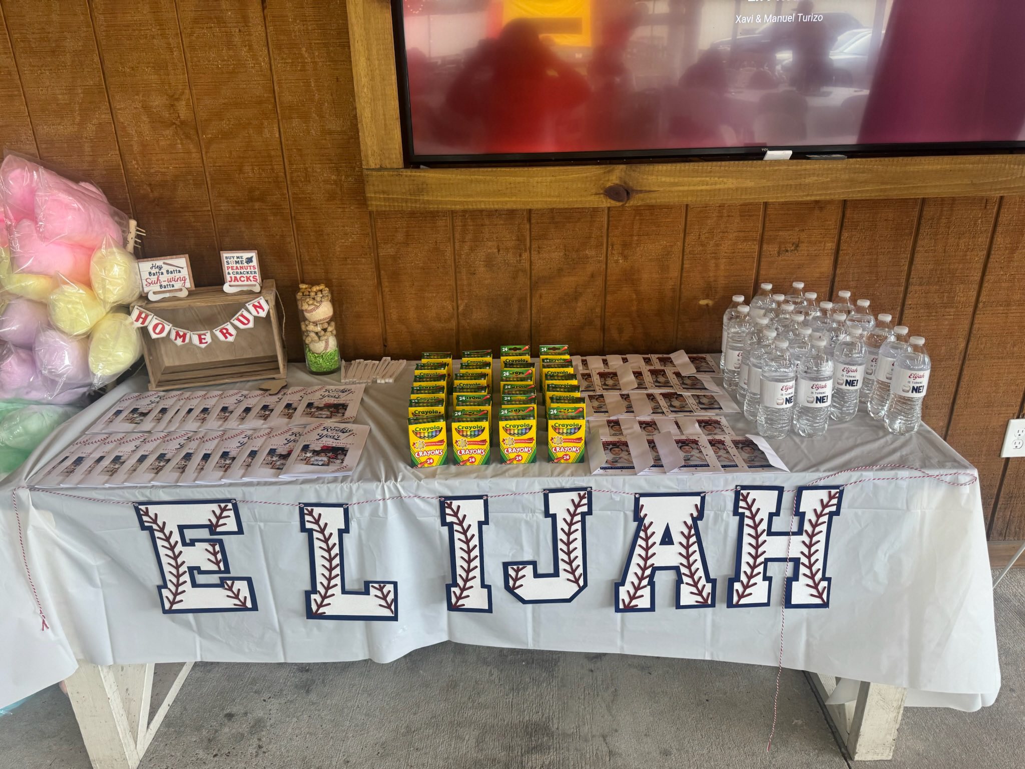 Baseball-themed birthday party table with cotton candy, coloring books, Crayola crayons, and water bottles labeled 'Elijah'.