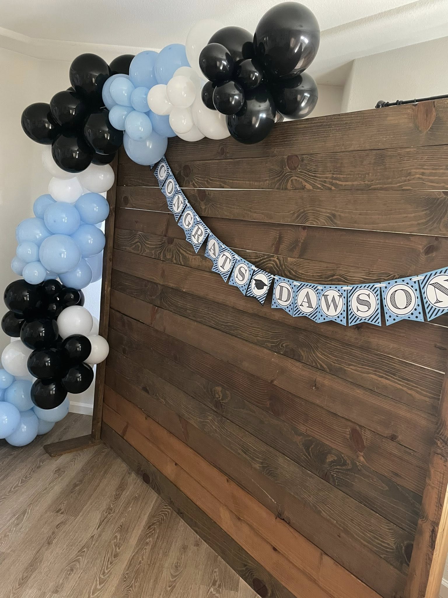 Rustic wood party backdrop featuring a Congratulations Dawson banner and a blue, black, and white balloon arch.