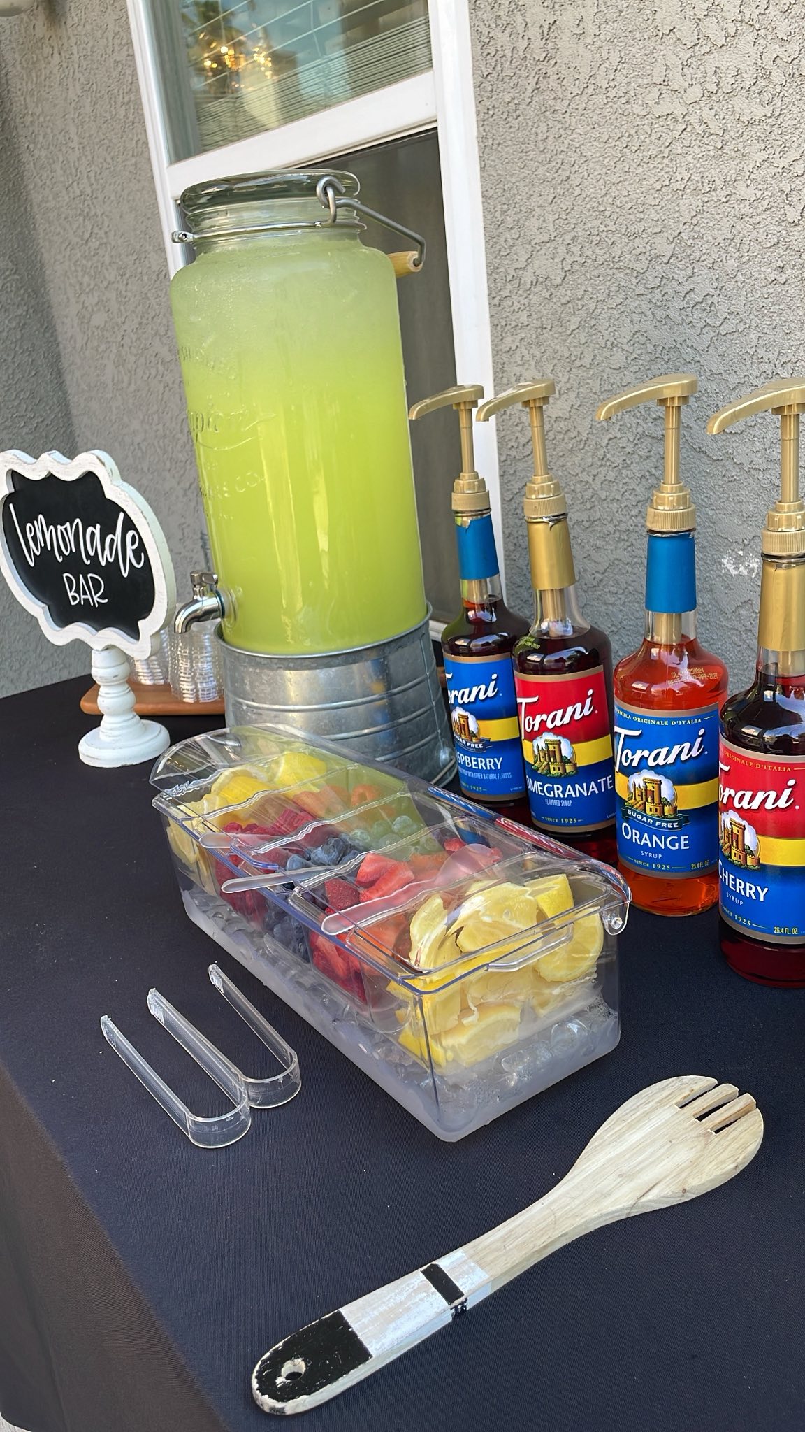 Lemonade bar setup with a large beverage dispenser, garnishes in an iced tray, and various Torani syrup pumps.