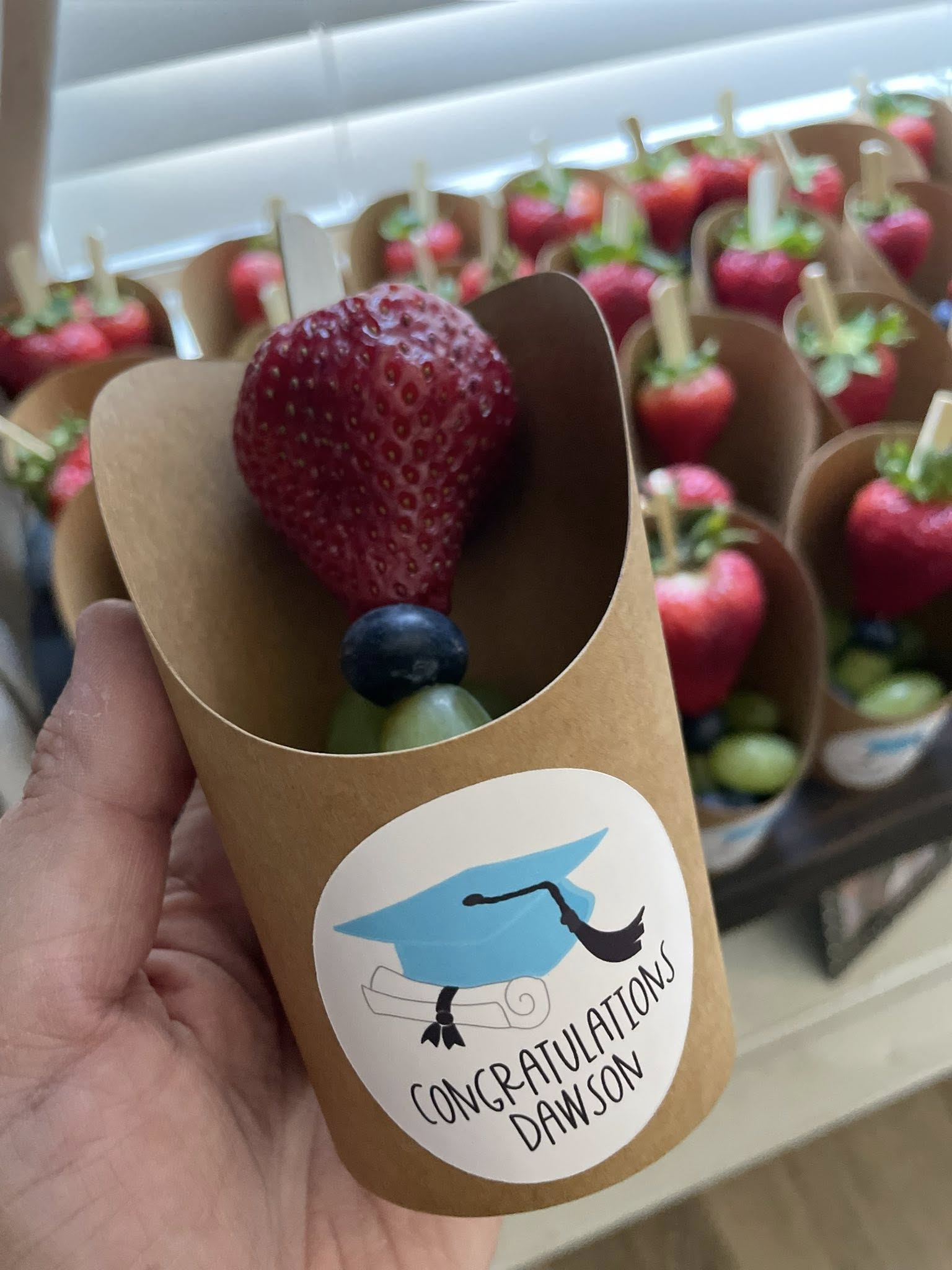 graduation-themed fruit cup with a strawberry, blueberry, and grape skewer in a paper holder with a mortarboard sticker