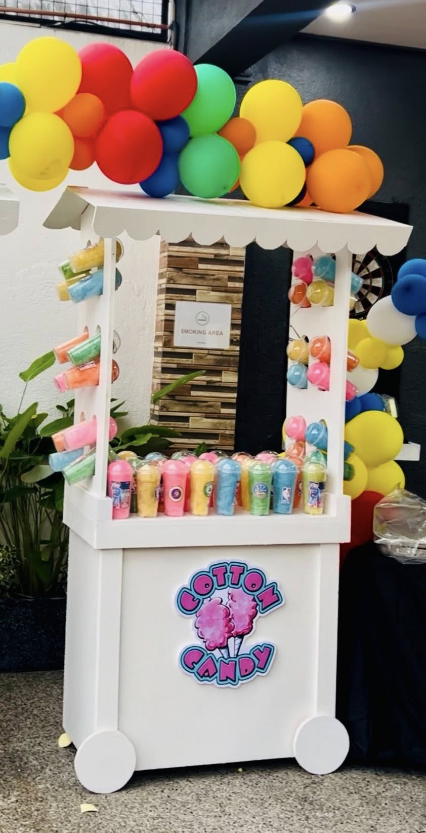 A white portable cotton candy stall with colorful balloons and displays of pre-packaged cotton candy cups.