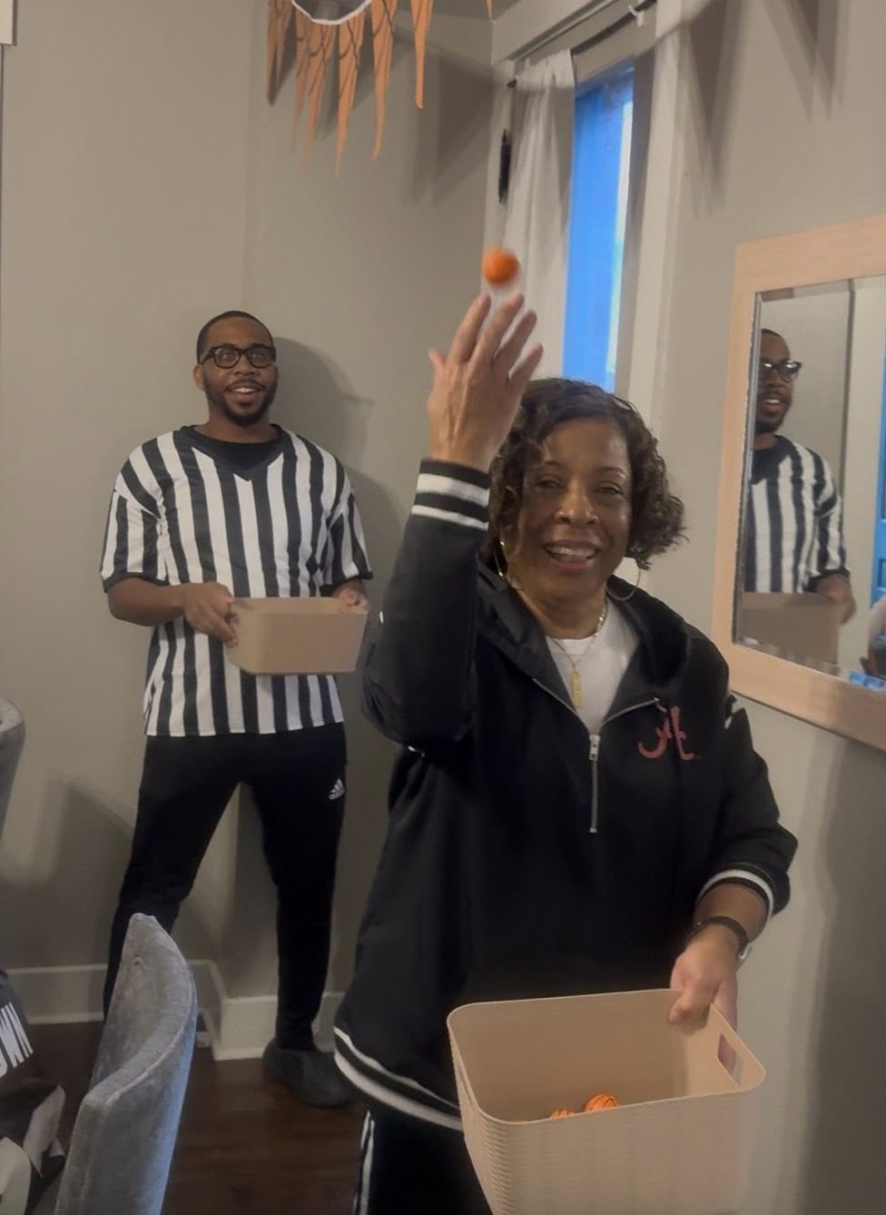 A woman tossing a small orange ball into a bin while a man in a referee costume watches in the background.