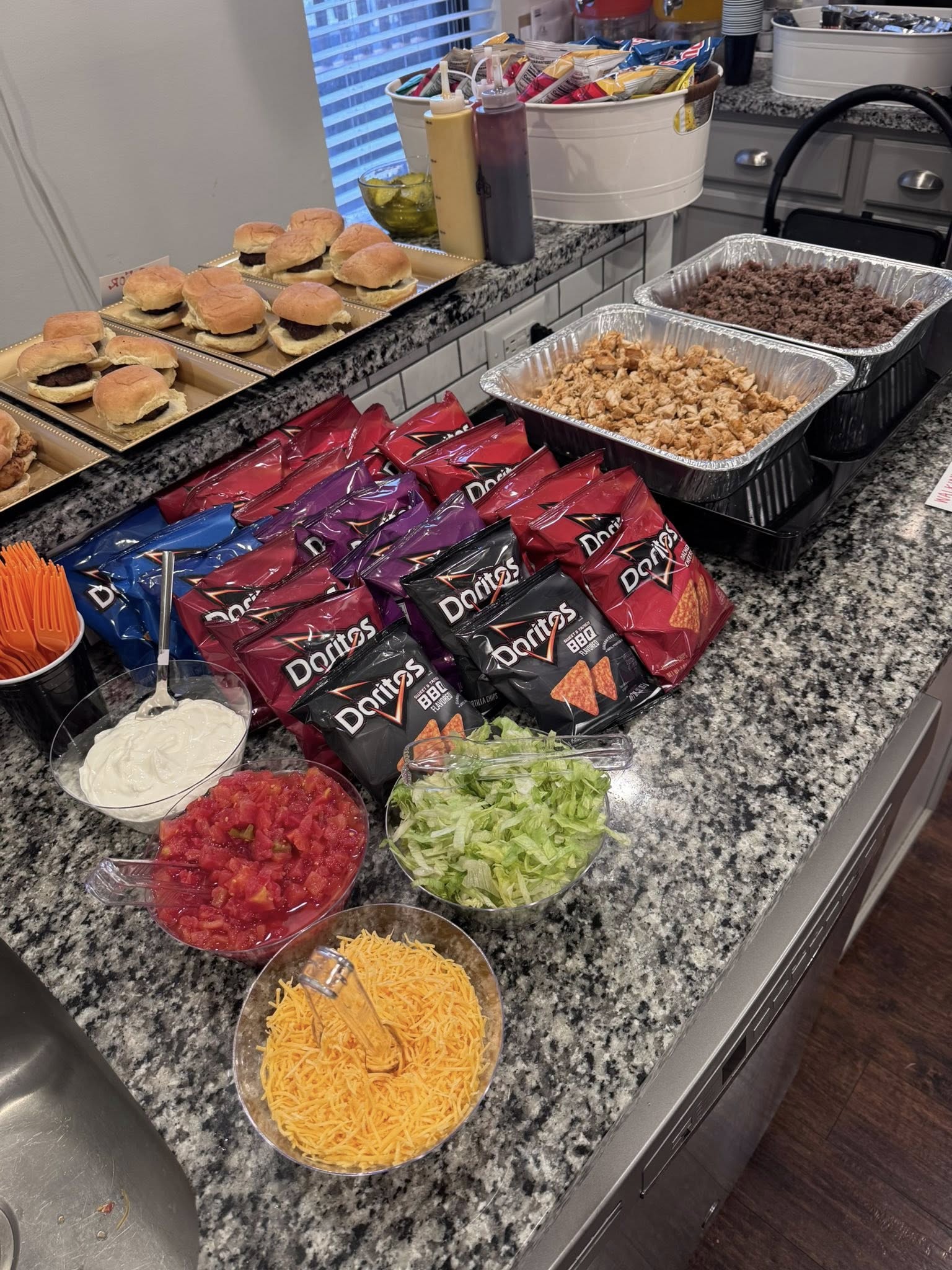 Catering table with slider burgers, seasoned meat, Doritos, and bowls of shredded cheese, lettuce, tomatoes, and salsa.