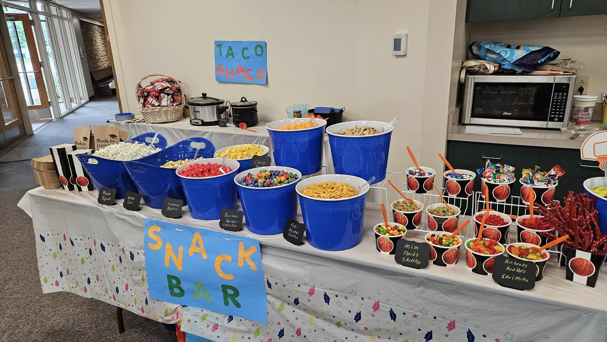 A self-serve snack bar with blue buckets of popcorn and small basketball-themed cups filled with assorted candies.