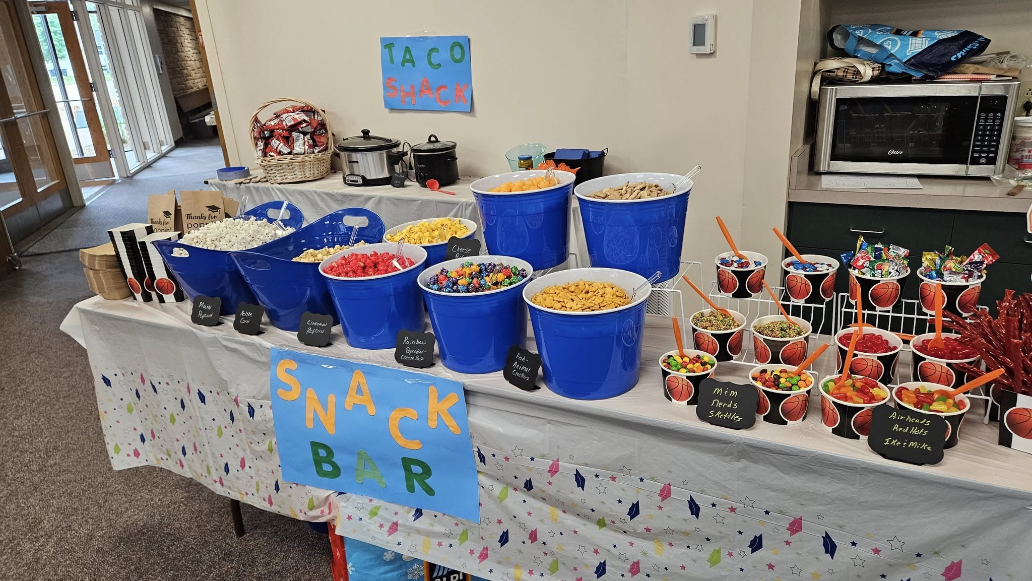 Snack bar setup featuring various bowls of popcorn, candy, and crackers arranged on a table with signs.