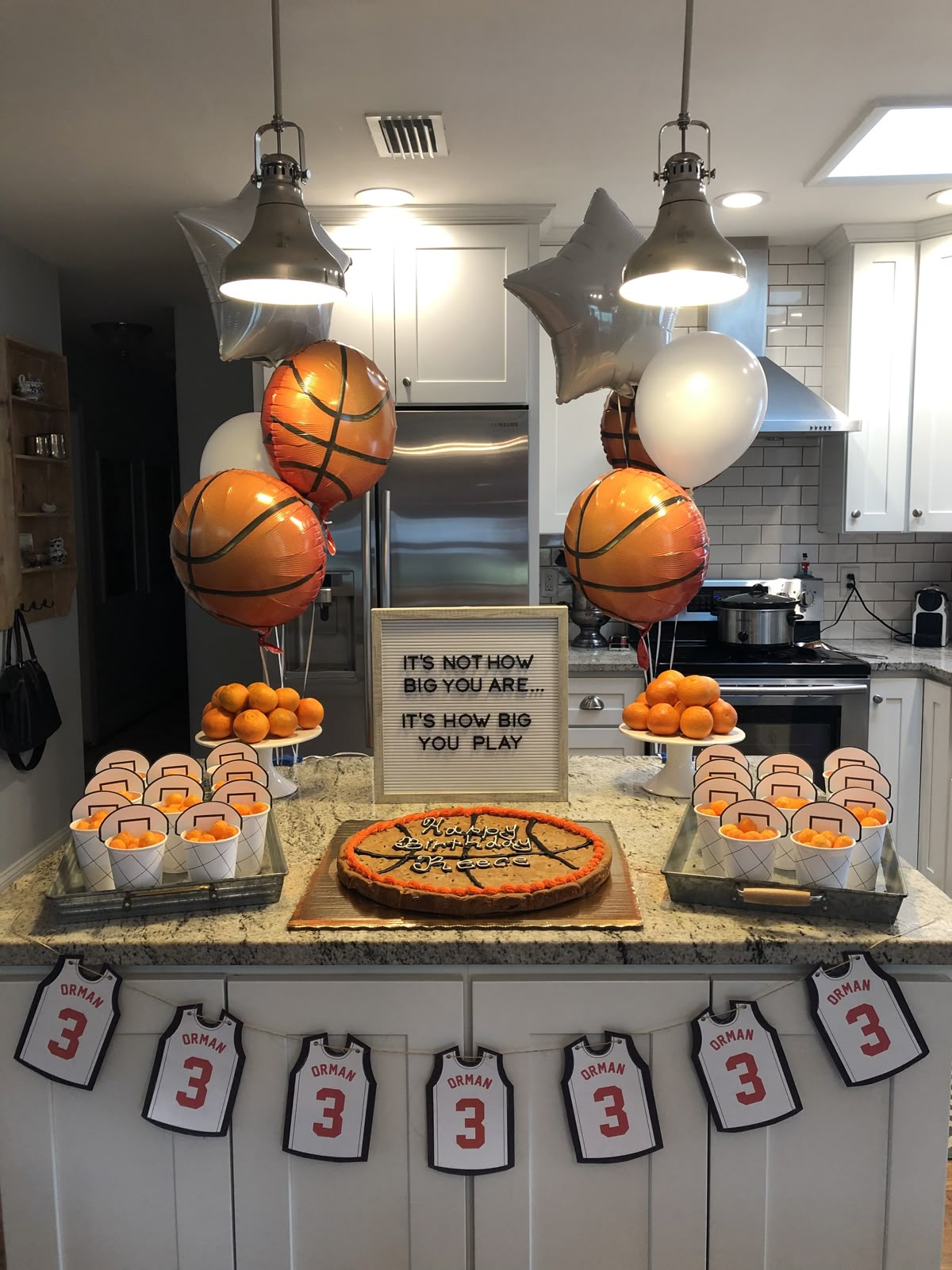 Basketball-themed birthday party setup with a cookie cake, basketball balloons, and snacks on a kitchen counter.