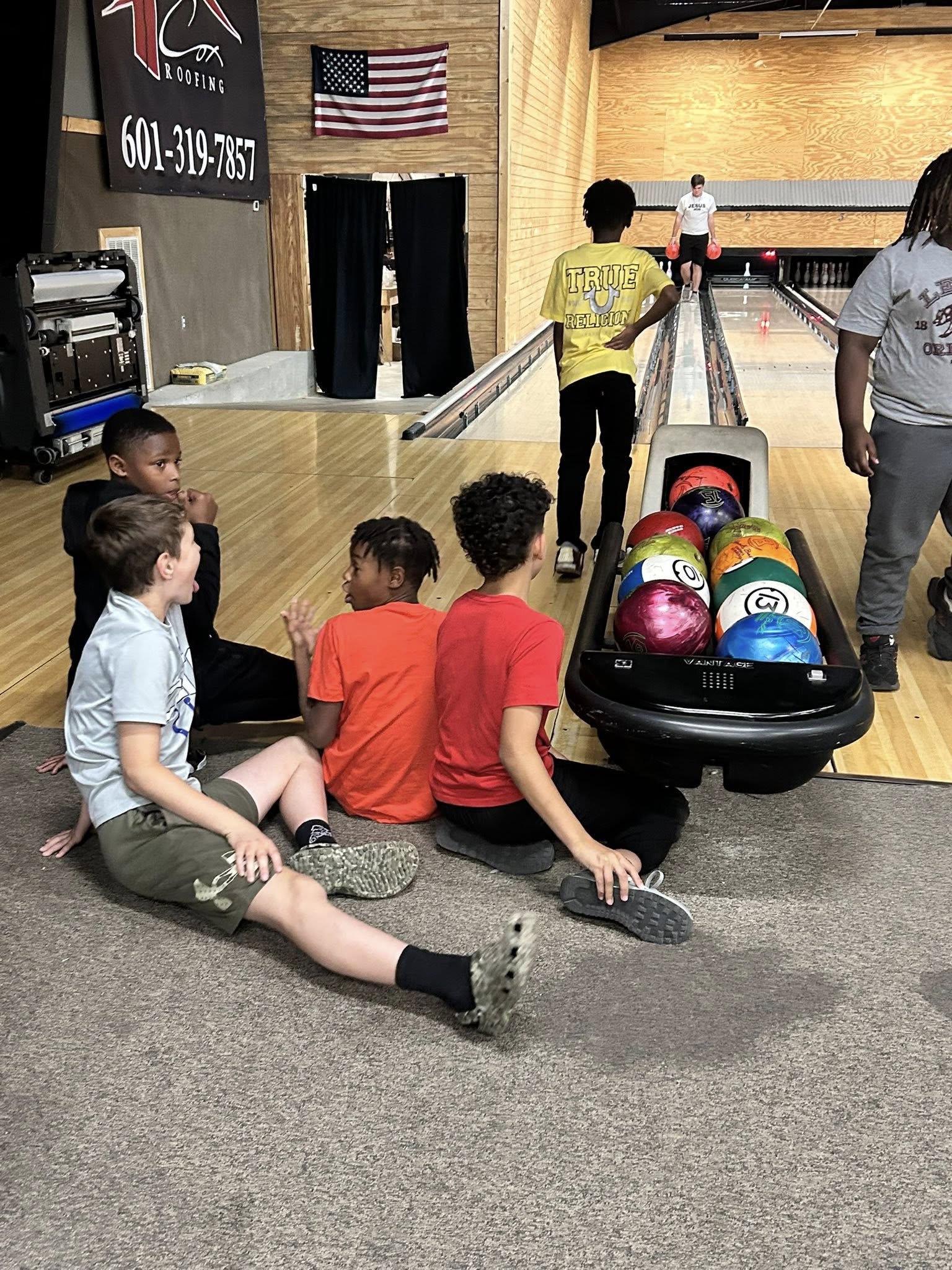 Group of young friends sitting on the floor behind a bowling ball return rack at a bowling alley.