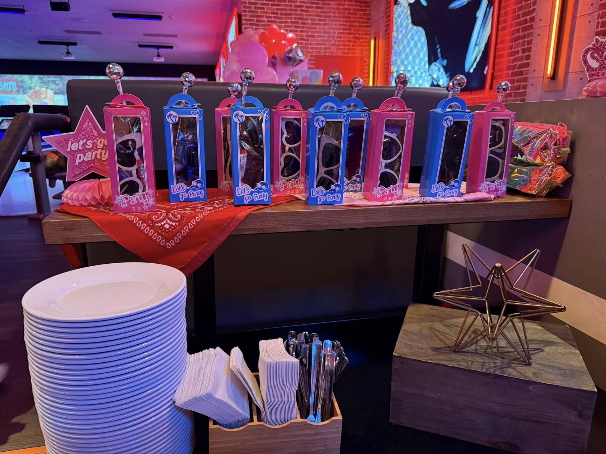 Table setup with rows of boxed Let's Go Party sunglasses, a stack of white plates, and party napkins.