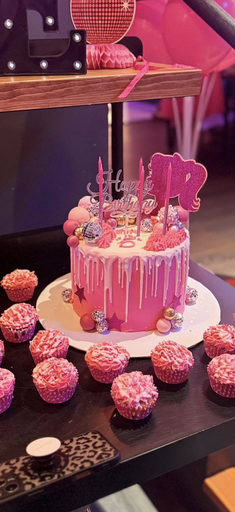 Pink birthday cake with white drip frosting, decorative disco balls, and a Barbie-themed cake topper.