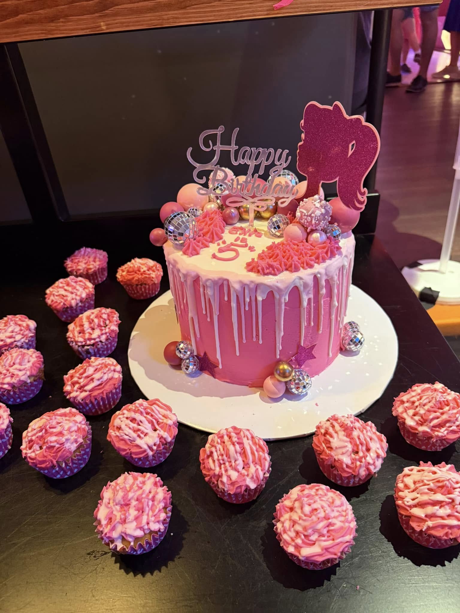 Pink birthday cake with white drip and disco ball decorations surrounded by matching frosted cupcakes.