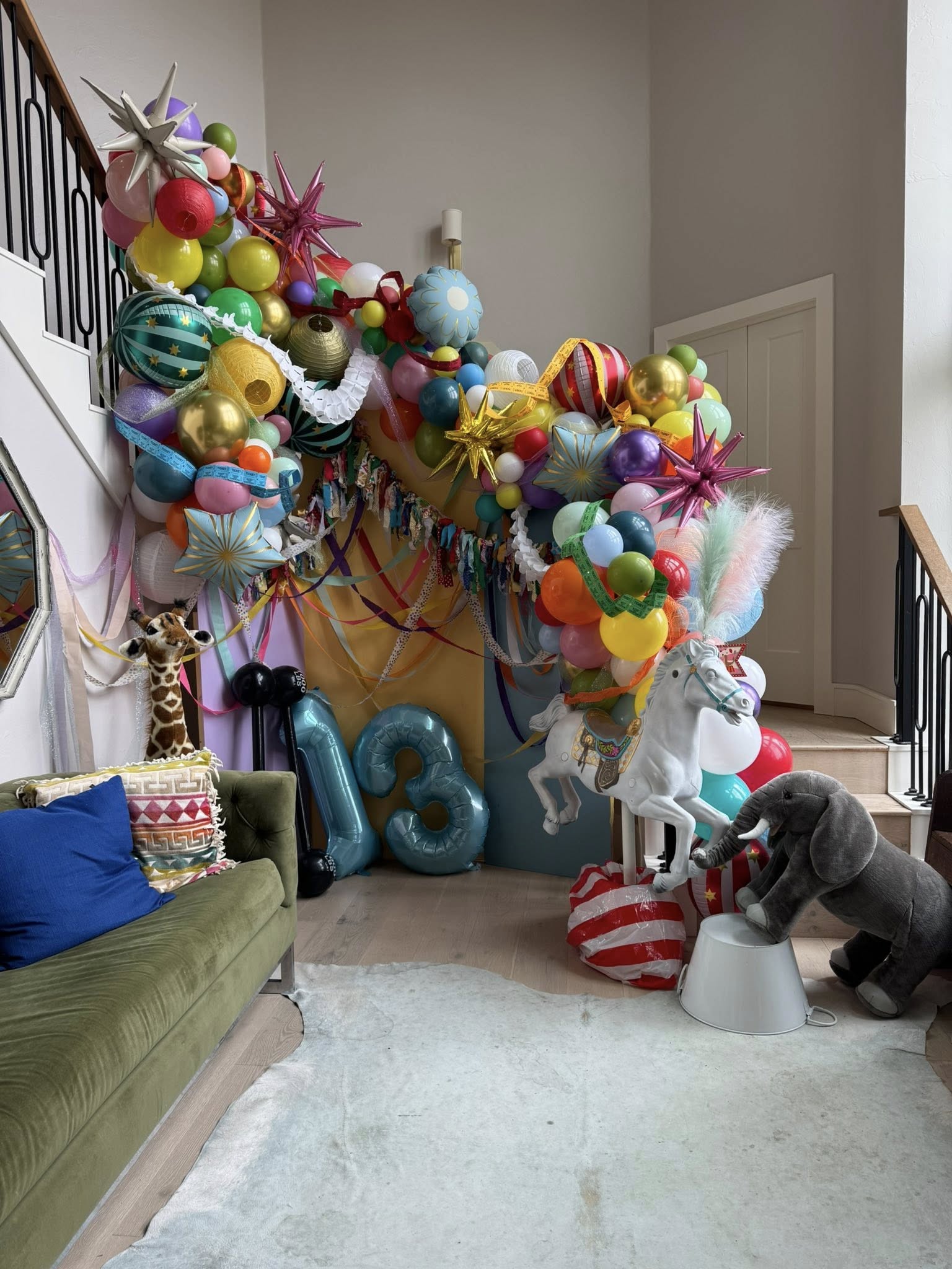 Colorful circus-themed birthday balloon installation with number 13 balloons and toy animals in a home entryway.