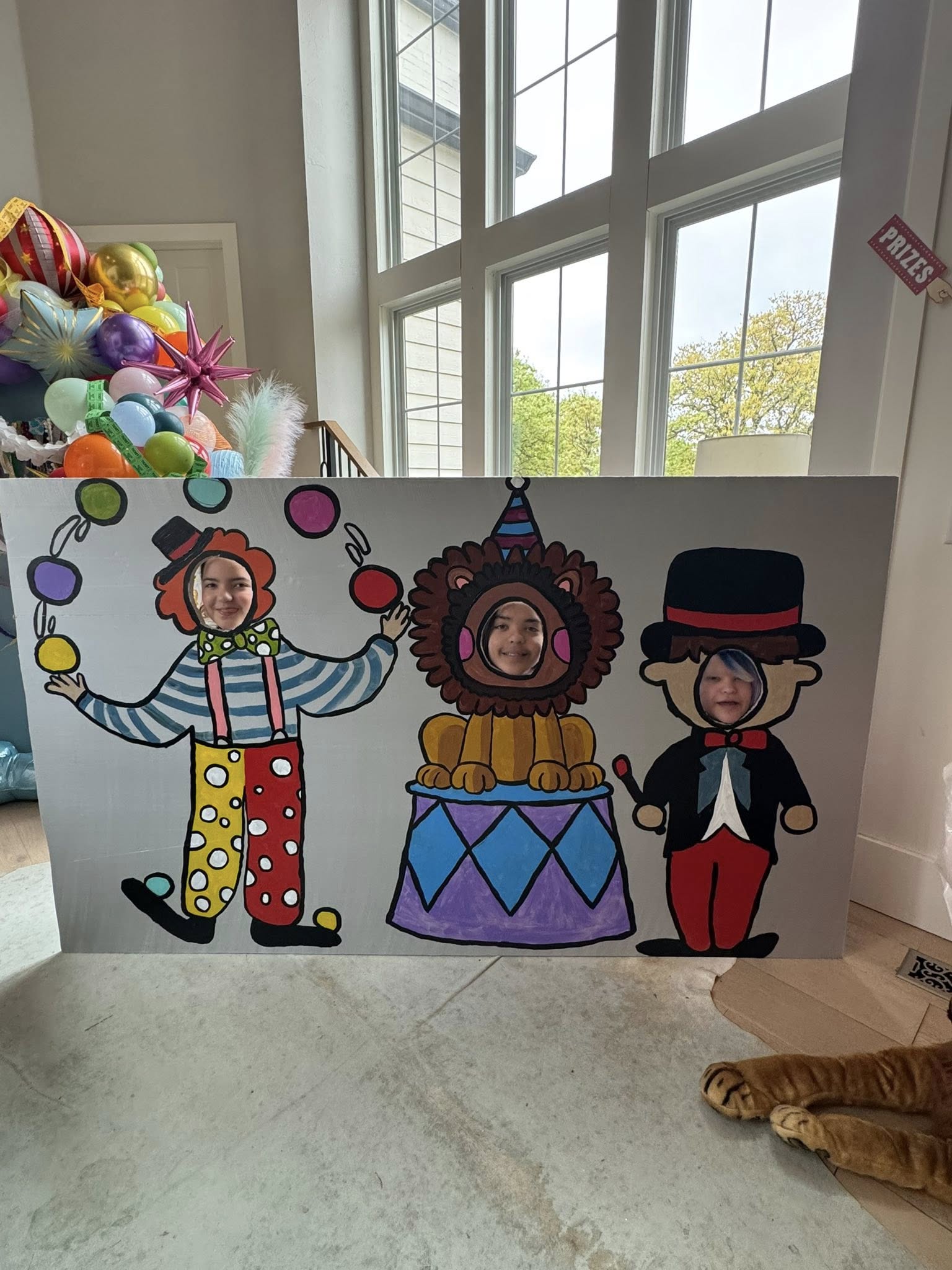 Three children posing behind a carnival-themed photo cutout board featuring a clown, a lion, and a ringmaster.