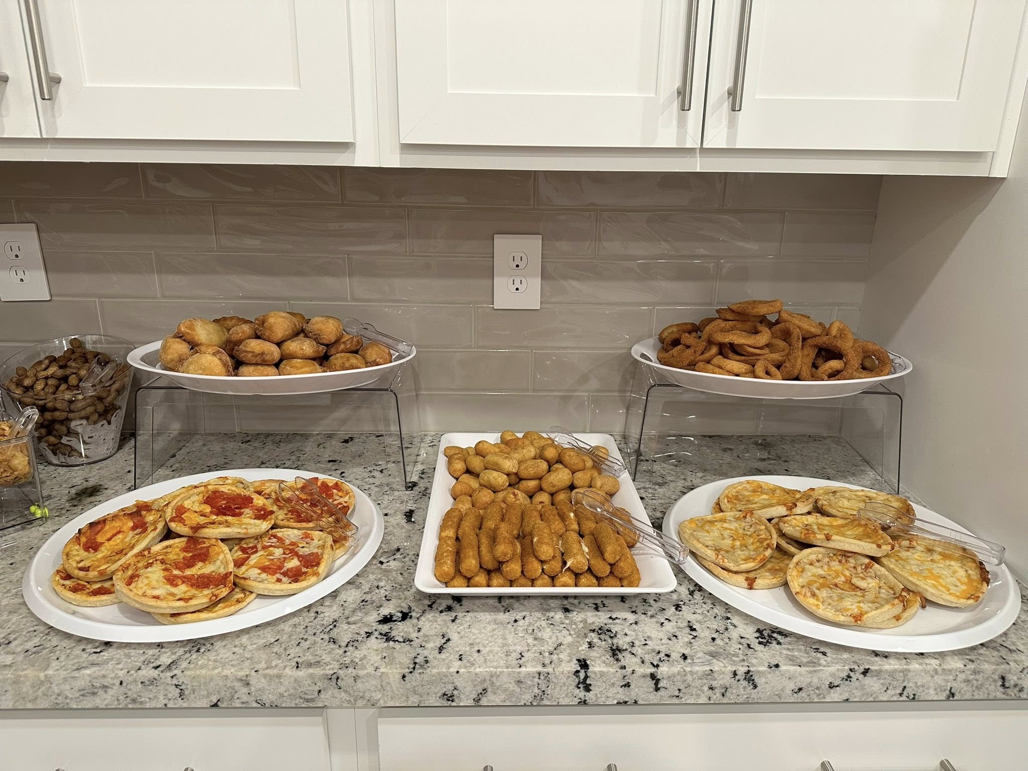 Catering spread with individual pizzas, mozzarella sticks, corn dogs, onion rings, and peanuts on a kitchen counter.