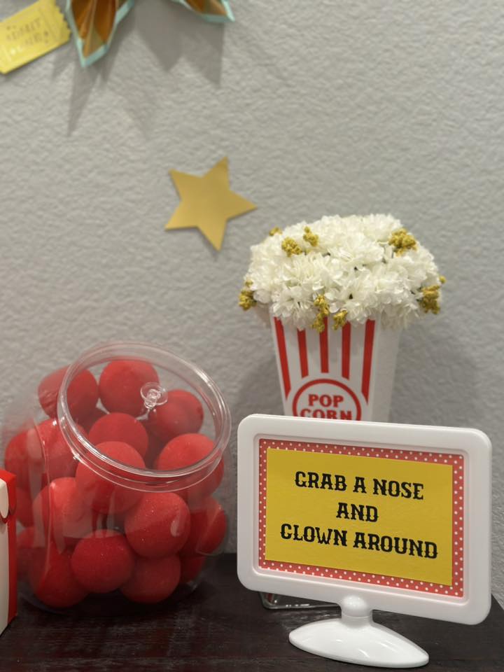 Table sign reading Grab a Nose and Clown Around next to a jar of clown noses and a popcorn bucket centerpiece.