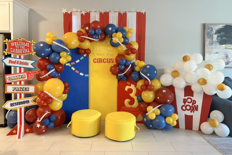Carnival-themed birthday party backdrop with colorful balloon arch, popcorn box display, and directional signs.