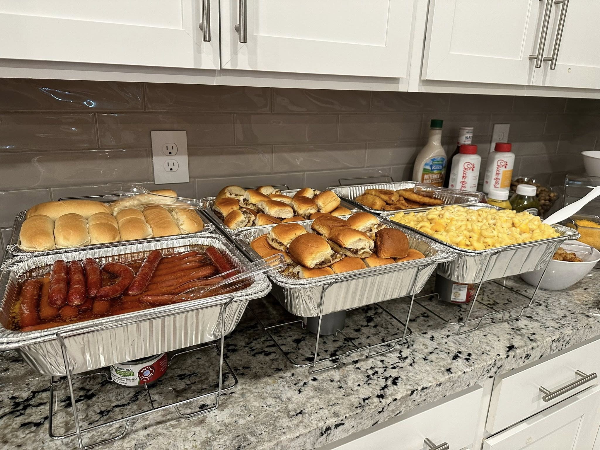 Catering trays filled with sausage, slider buns, mini sandwiches, mac and cheese, and chicken tenders.
