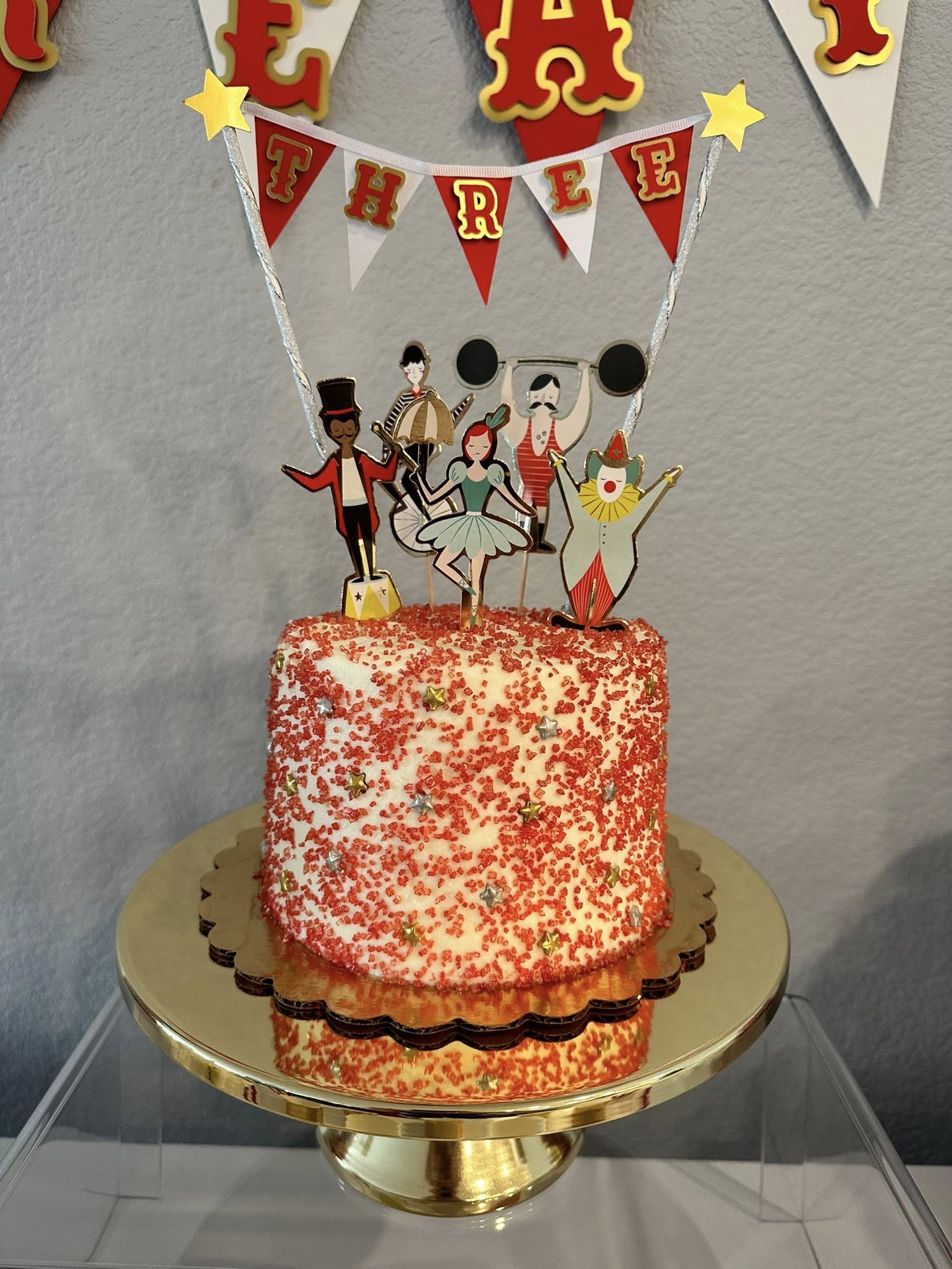Cake decorated with red sprinkles and a circus-themed topper featuring a ringmaster, ballerina, and clown.