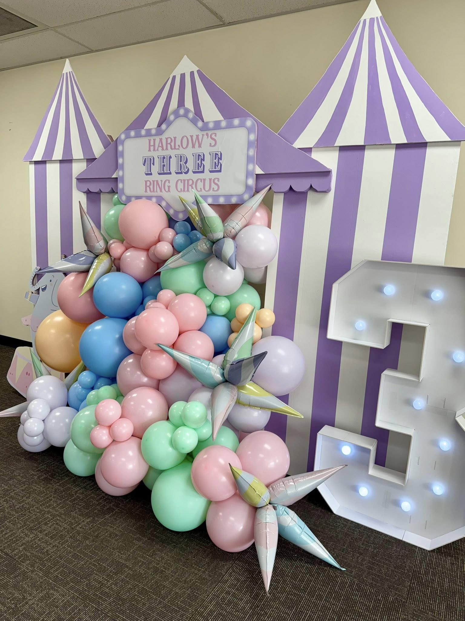 Circus themed birthday party backdrop for a three-year-old with pastel balloon arch and illuminated number three.